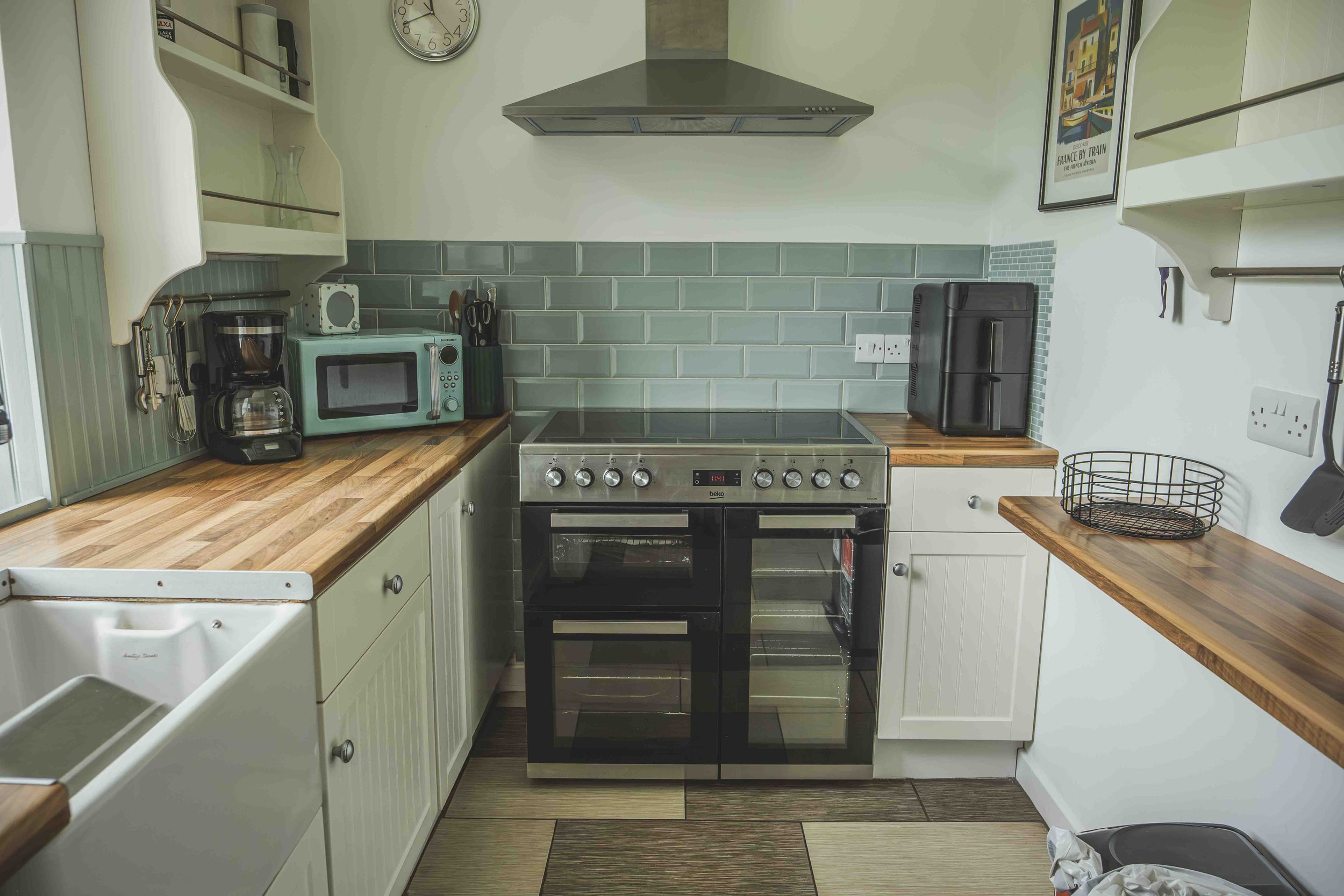 A small kitchen with wooden countertops, white cabinets, a black oven with a stovetop, a microwave, a coffee maker, and various utensils against a tiled backsplash. There are wall-mounted shelves, a clock, and a framed poster.