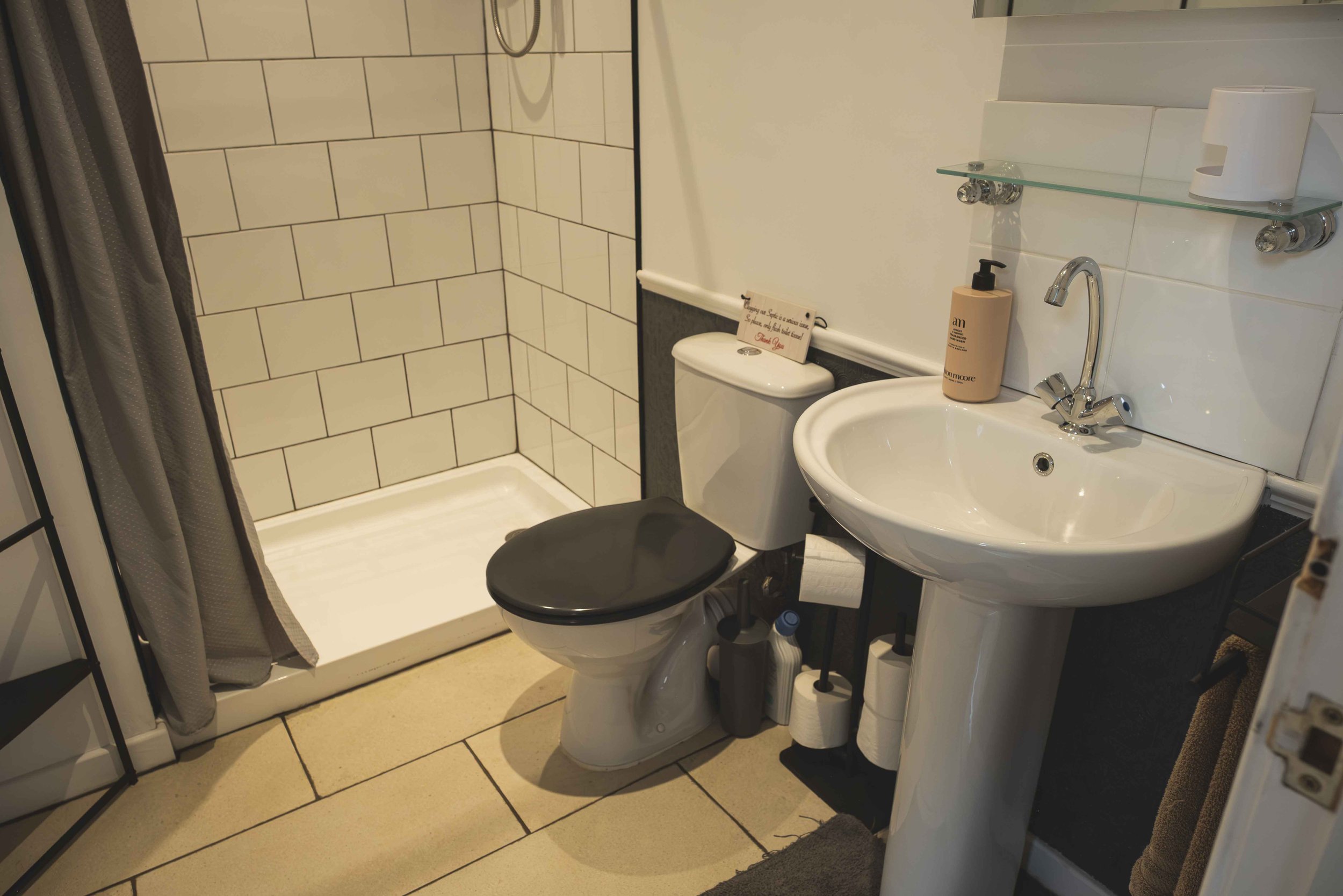 A small bathroom with a shower area that has a curtain, a toilet, and a sink with a mirror and shelves. There is a bottle of hand soap on the sink and a roll of toilet paper on the shelf.