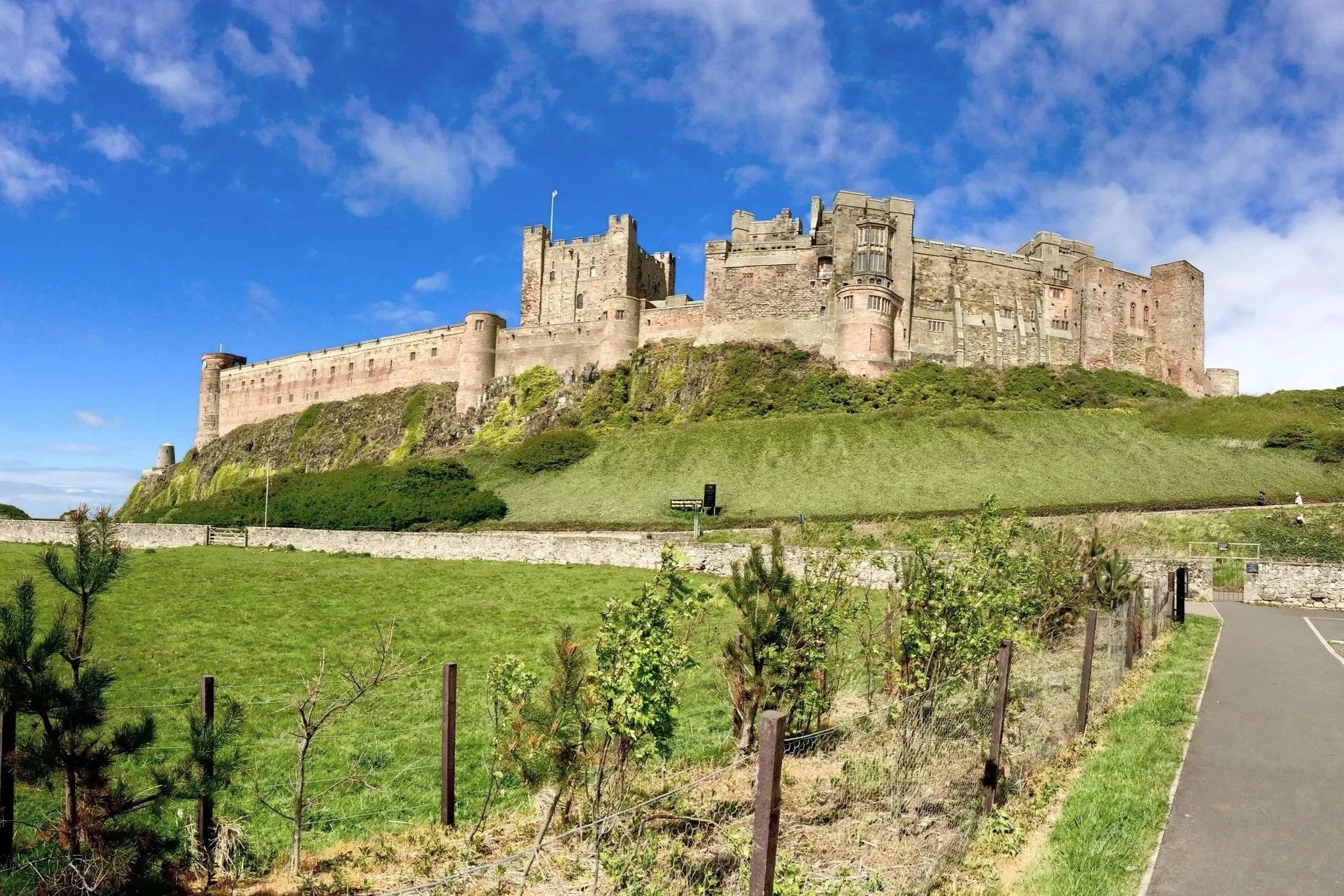 A large medieval castle atop a grassy hill with a clear blue sky and some clouds in the background.