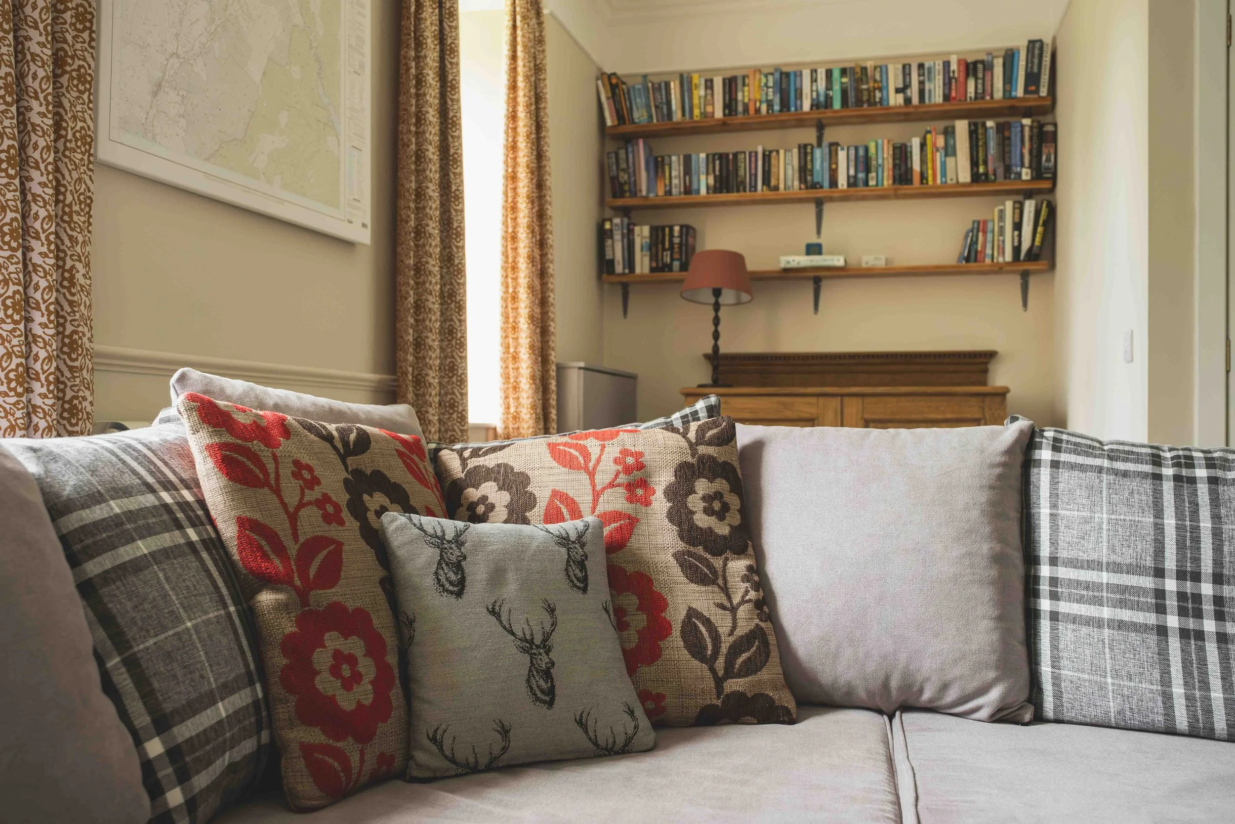 A cozy living room corner with a sofa covered in various patterned throw pillows, a window with floral curtains, and a set of wooden shelves filled with books on the wall.