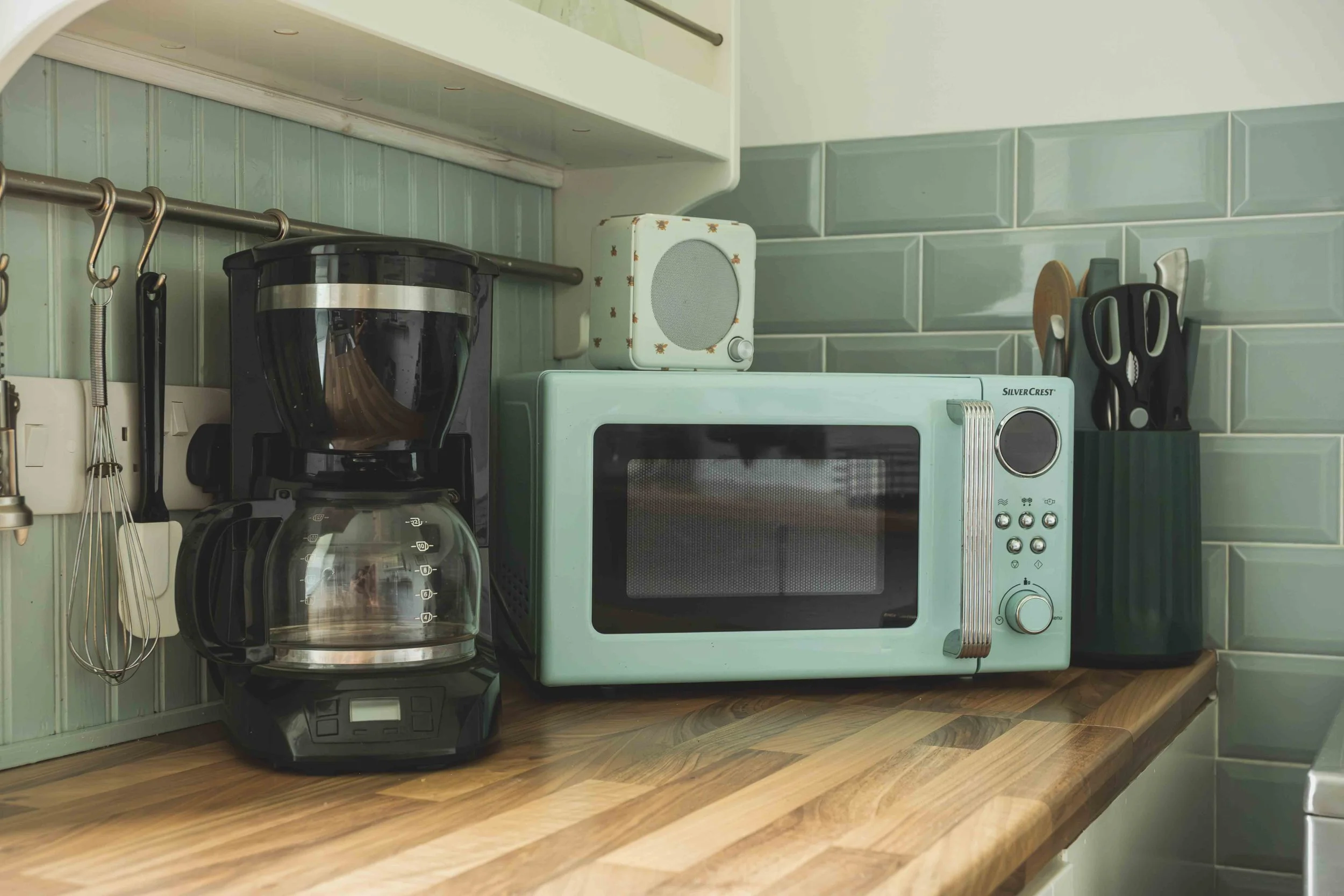 Kitchen countertop with a black coffee maker, a teal microwave, a small white speaker with a floral pattern, and kitchen utensils on a green tiled wall behind.