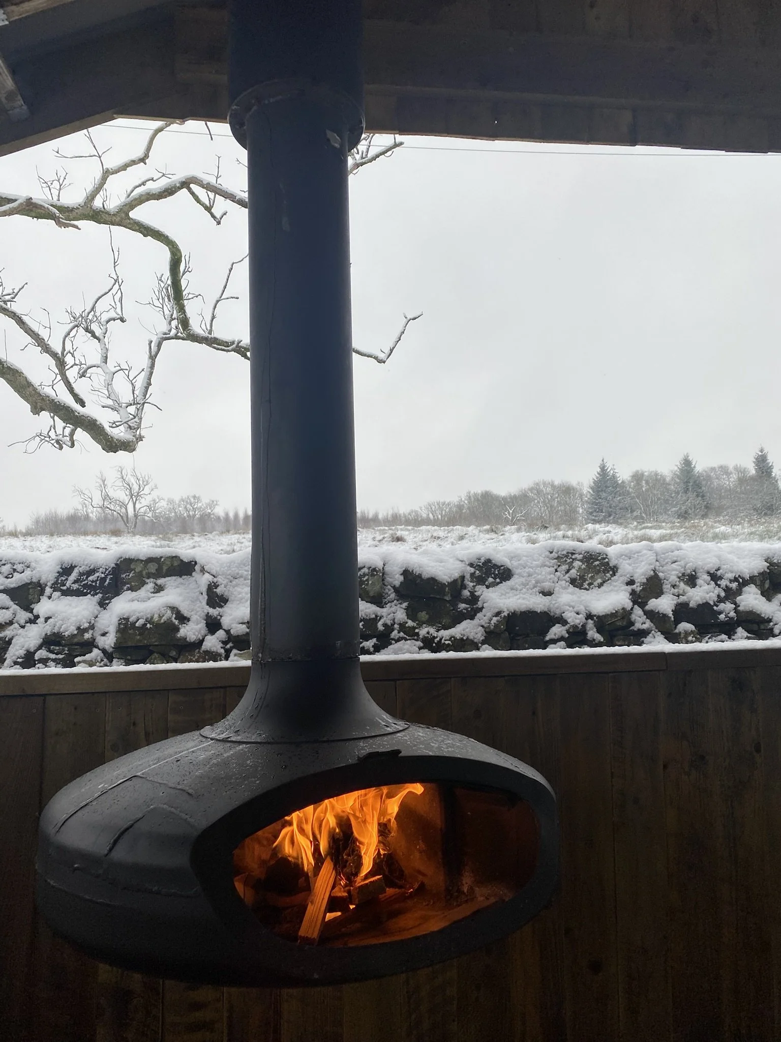 A wood-burning stove with a fire inside, viewed from the interior of a porch or balcony. Snow covers the ground and trees are visible in the background, and the stove pipe extends upwards through the ceiling.