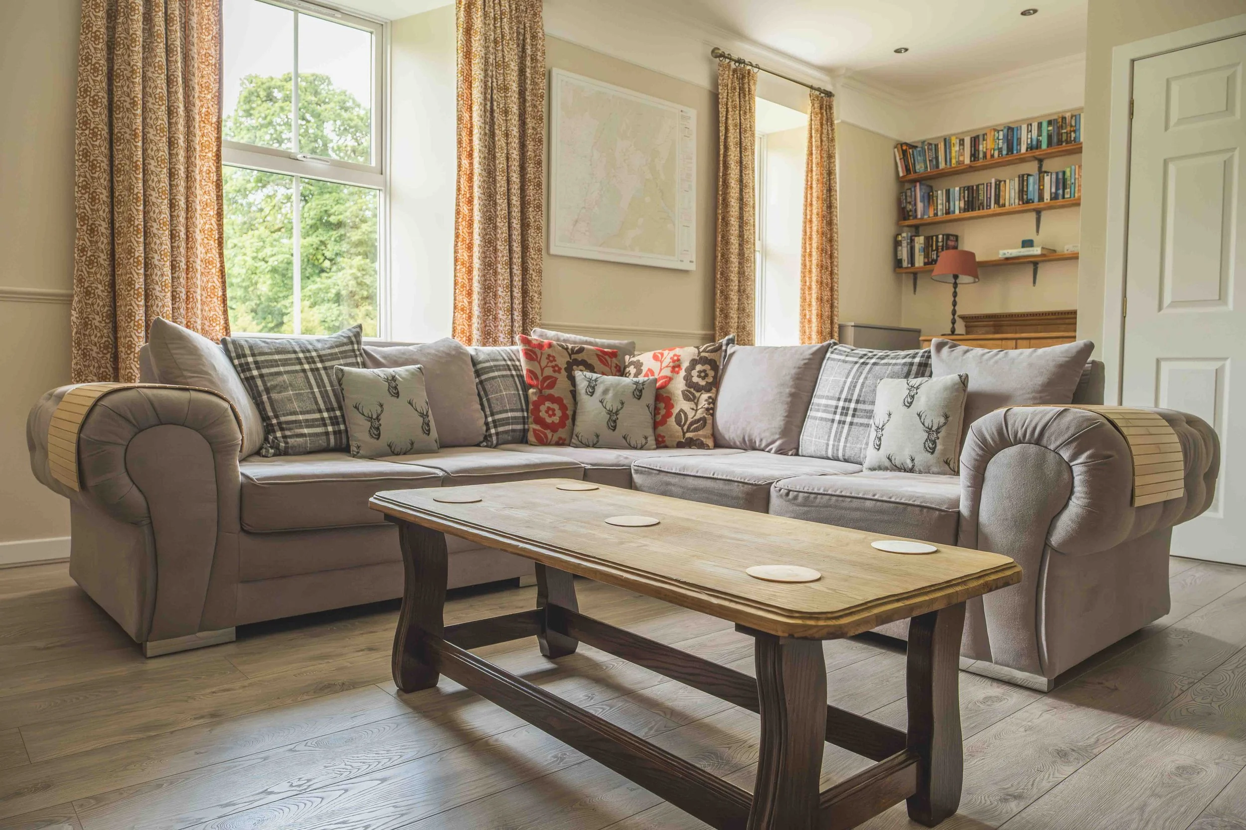 Living room with large gray sectional sofa decorated with plaid, floral, and deer motif pillows, next to a rustic wooden coffee table, large windows with patterned curtains, a map on the wall, and a bookcase with books and a lamp.