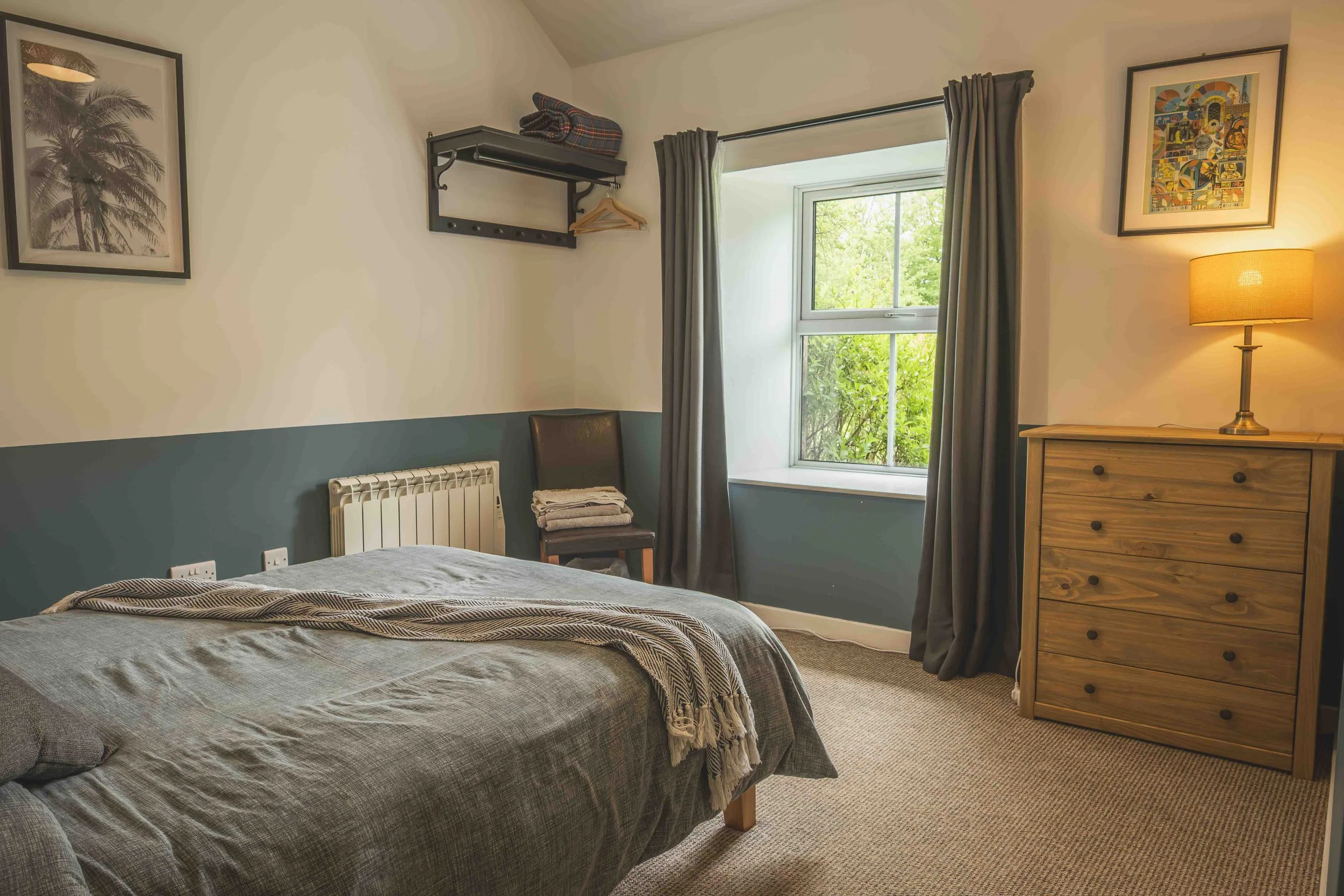 A cozy bedroom with a bed covered in a gray blanket and pillows, a wooden dresser with a lamp, a window with gray curtains showing greenery outside, framed artwork on the walls, a chair with folded towels, and a wall shelf with folded blankets and ha