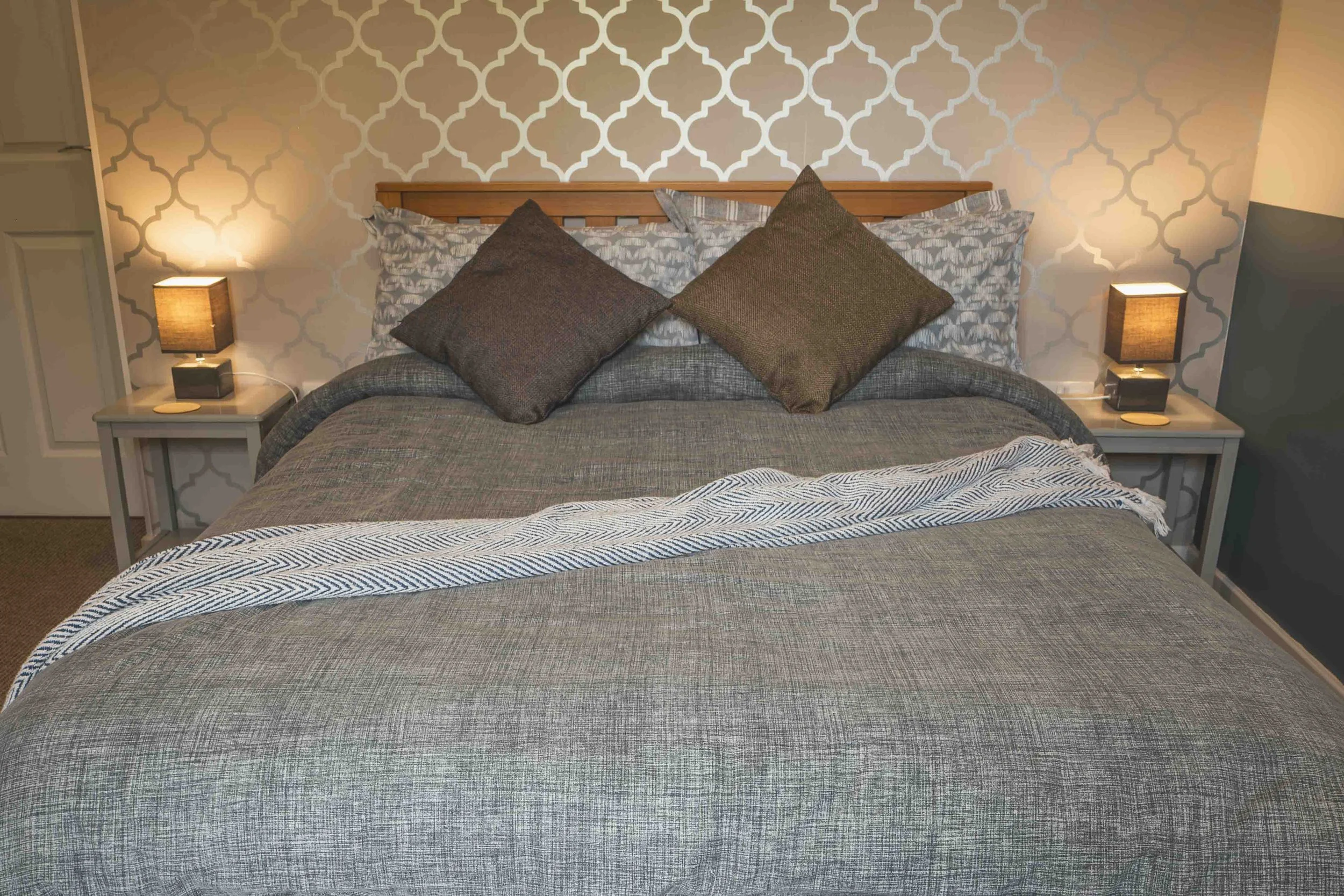 A neatly made king-sized bed with a gray and brown textured bedspread, accented with three large pillows in brown covers and two standard pillows with a leaf pattern, set against a beige wall with geometric wallpaper. There are matching bedside table