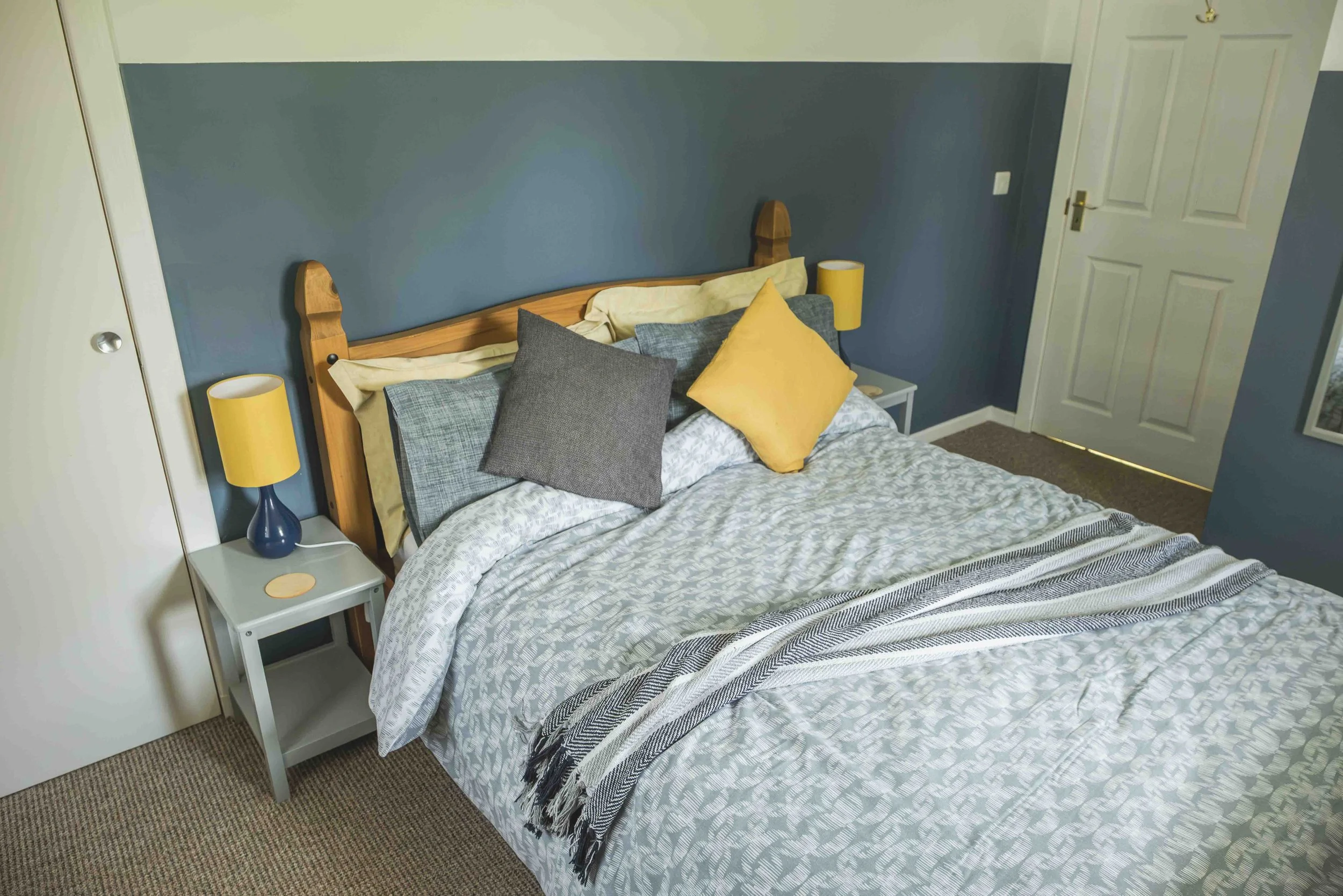 A neatly made bed with patterned comforter and multiple pillows, flanked by two yellow bedside lamps on white nightstands, in a room with a blue accent wall, white closet, and door.