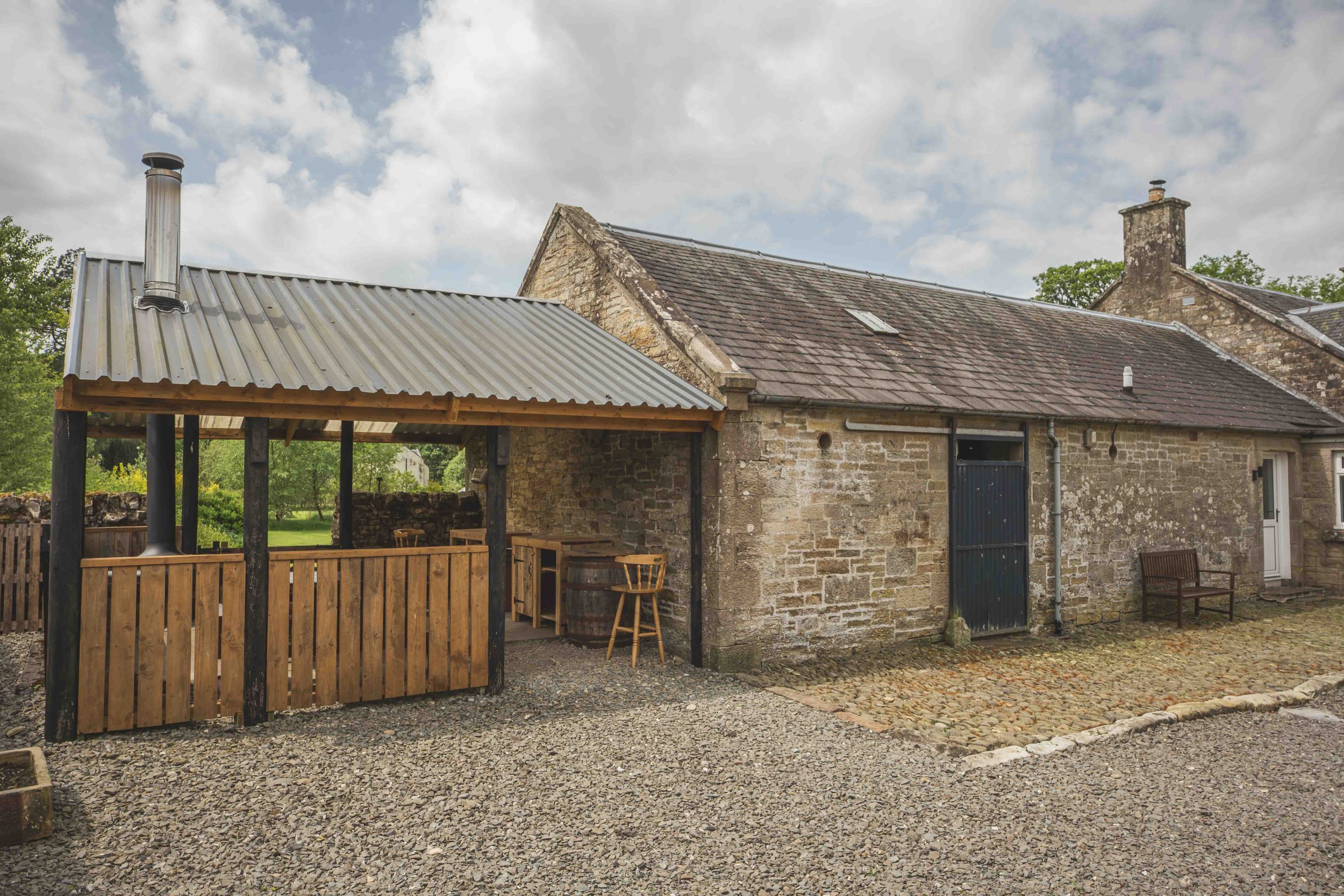 A stone building with a sloped roof, chimney, and window under partly cloudy sky. An attached wooden and metal covered outdoor seating area with chairs, barrels, and a fence surrounds a gravel ground.