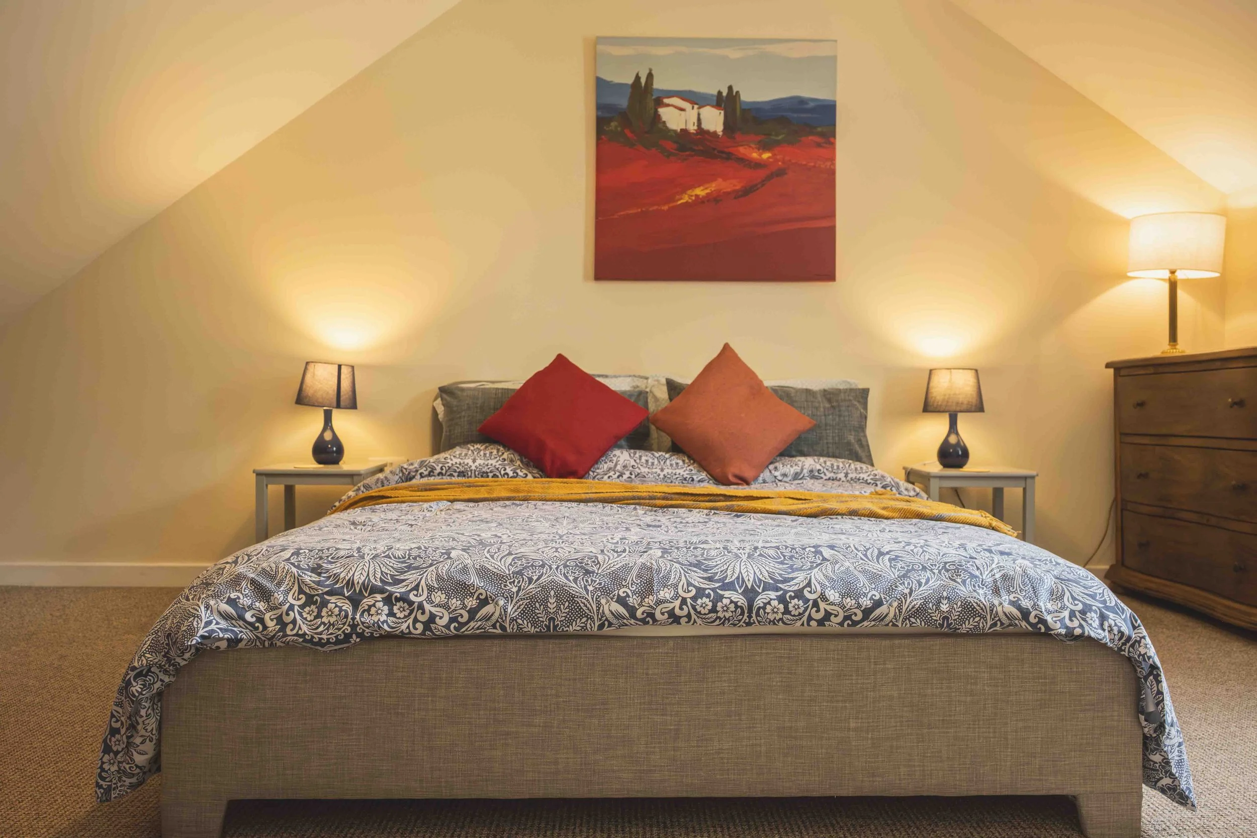 A neatly made bed with a patterned blue and white bedspread, two red throw pillows, and a yellow blanket, flanked by two bedside tables with black lamps. A landscape painting hangs on the yellow wall above the bed, and a tall dresser with a lamp is o