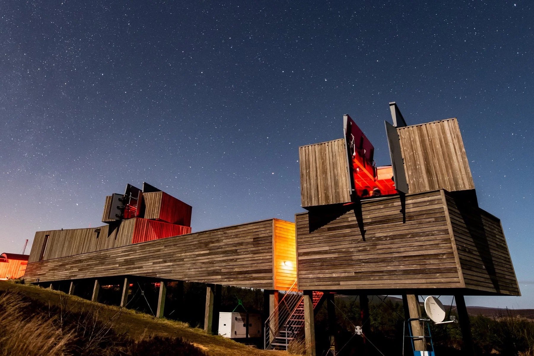 Nighttime view of a modern house with wooden exterior, elevated on stilts, set against a starry sky. Some windows emit a red glow.