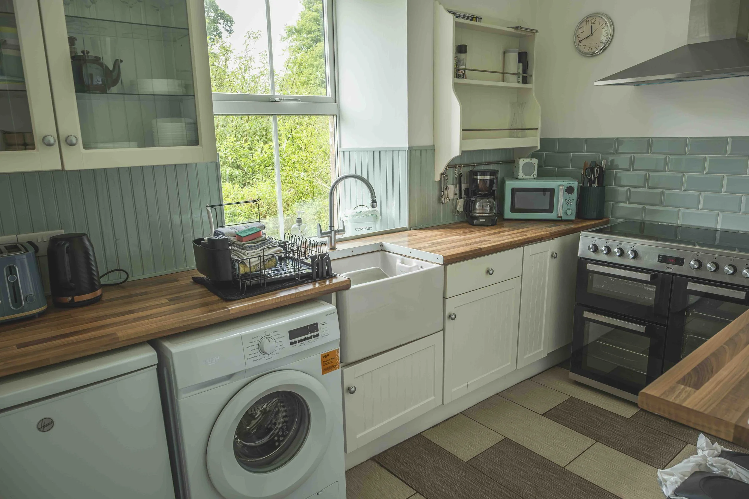 A kitchen with green tiled walls, a wooden countertop, a white sink, a microwave, a coffee maker, a toaster, a washing machine, and a gas stove. There is a window with a view of greenery outside, and various kitchen utensils and appliances are placed