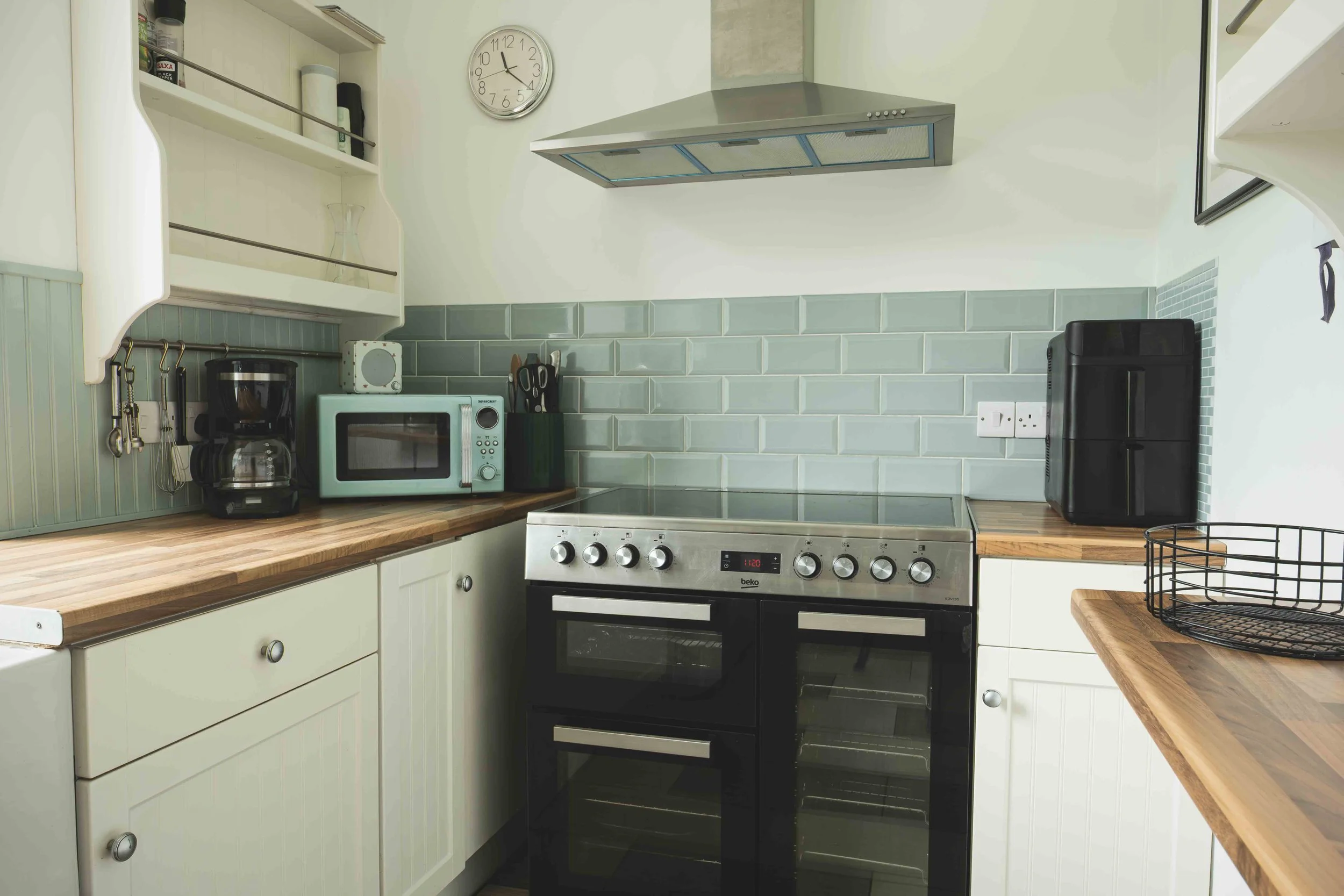 A modern kitchen with white cabinets, a wooden countertop, a stainless steel stove, a microwave, a coffee maker, and a black dishwasher. There is a wall clock and a small shelf with various items, including a glass. There is also a black wire basket 