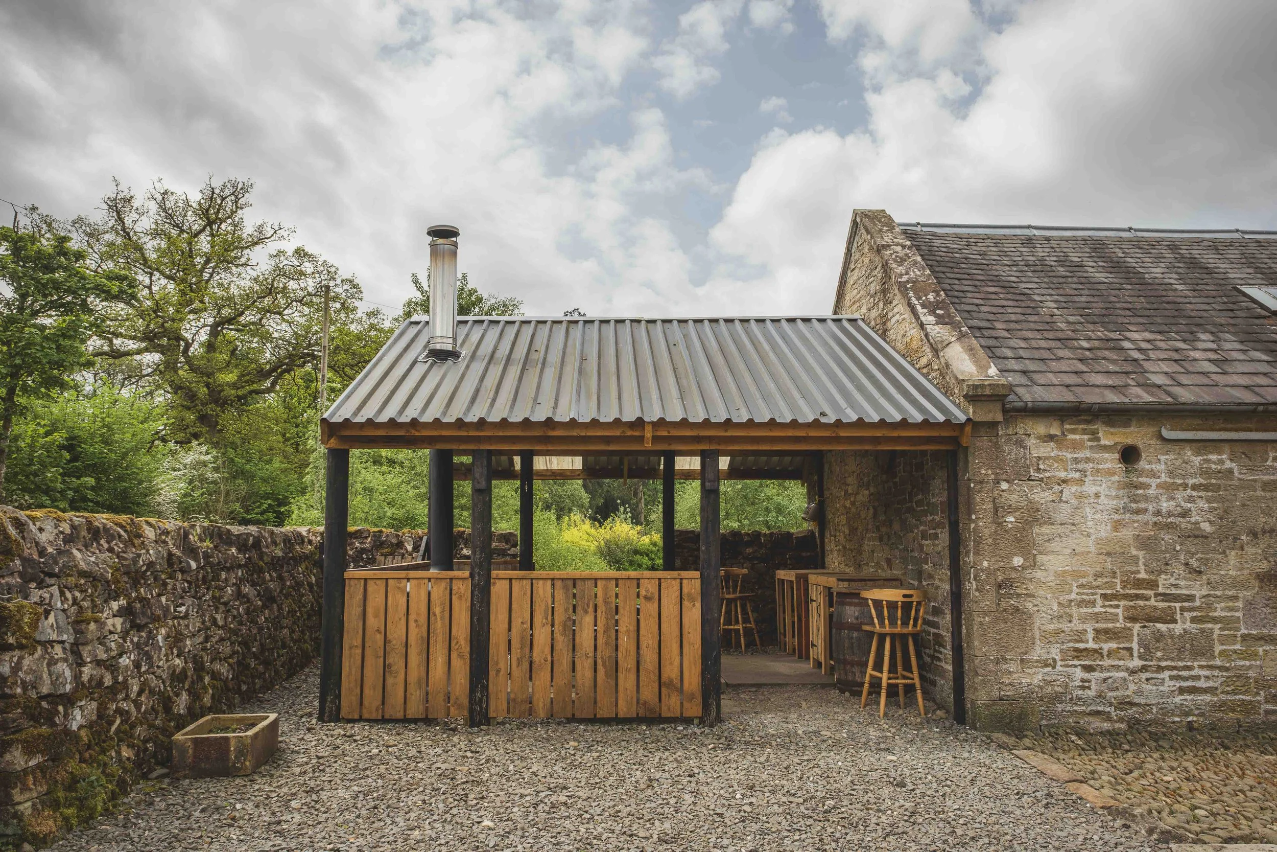 A rustic outdoor patio area with a wooden bar and high chairs in front of a stone building with a sloped roof, surrounded by trees and a stone wall, under cloudy sky.