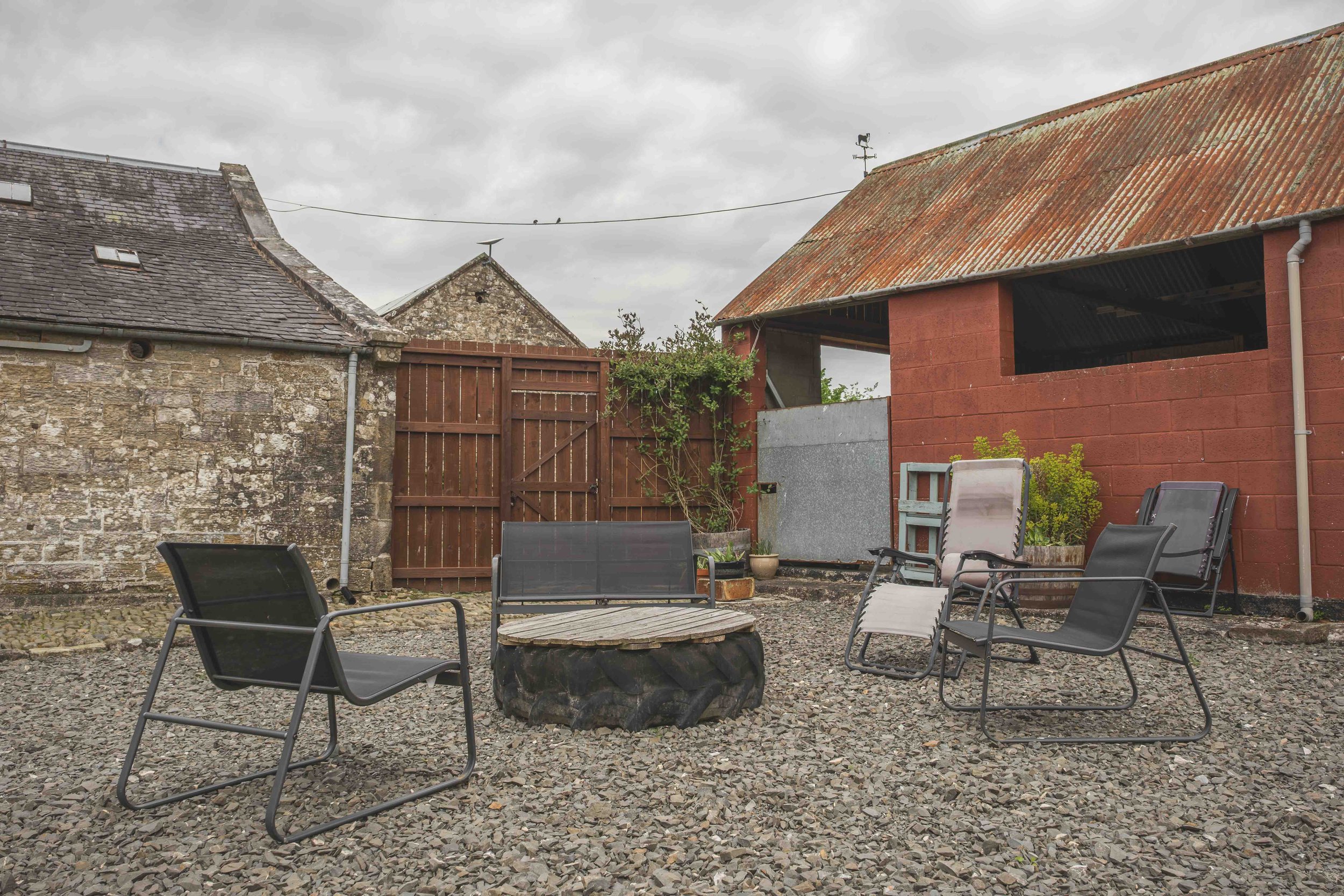 A backyard patio area with multiple black chairs around a circular fire pit made of stones, with a stone and brick house and a wooden shed with a rusted metal roof under an overcast sky.
