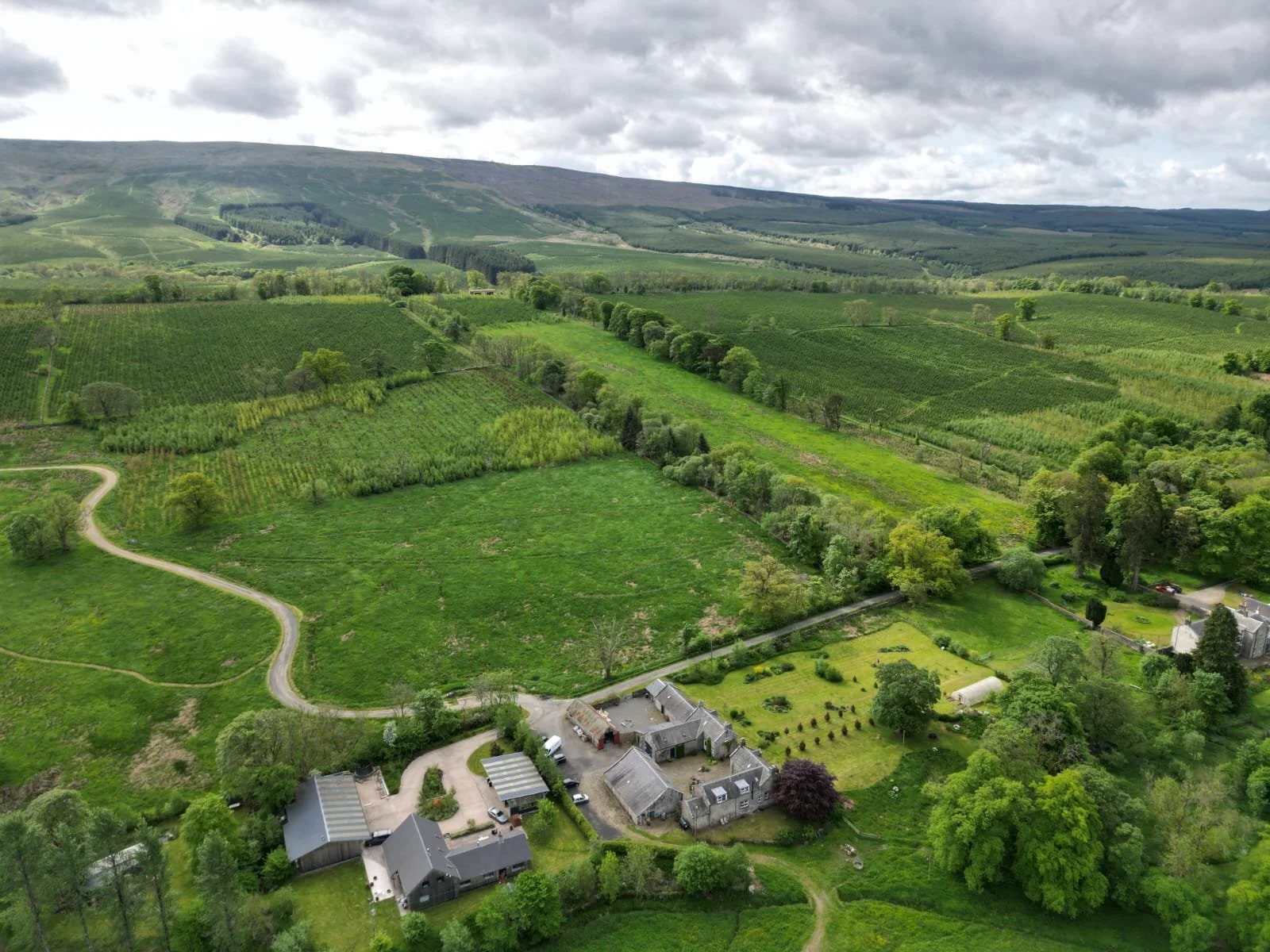 Aerial view of a lush green countryside with rolling hills, farmland, trees, a winding road, and a few houses and buildings.