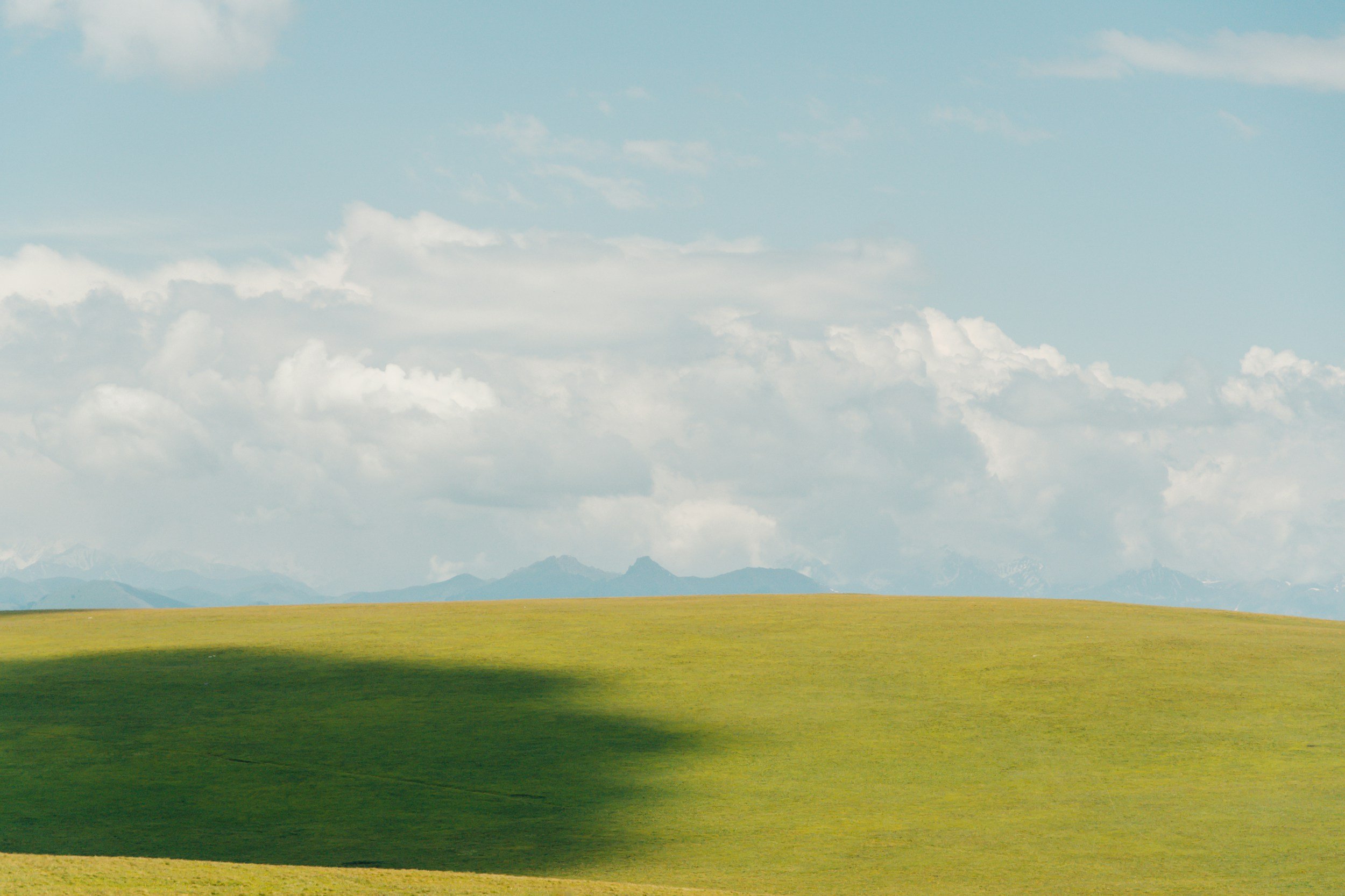 Open grassy field with a large shadow cast on it, under a partly cloudy sky with distant mountains.