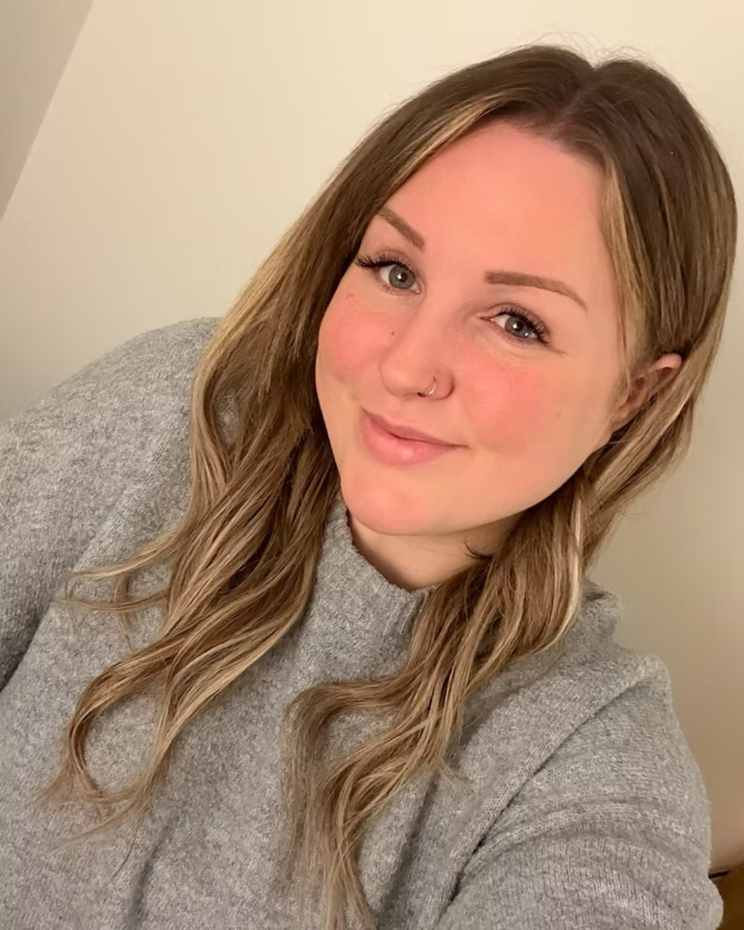 A woman with long, wavy, light blonde hair and blue eyes, wearing a gray sweater, taking a selfie indoors with a plain beige wall in the background.