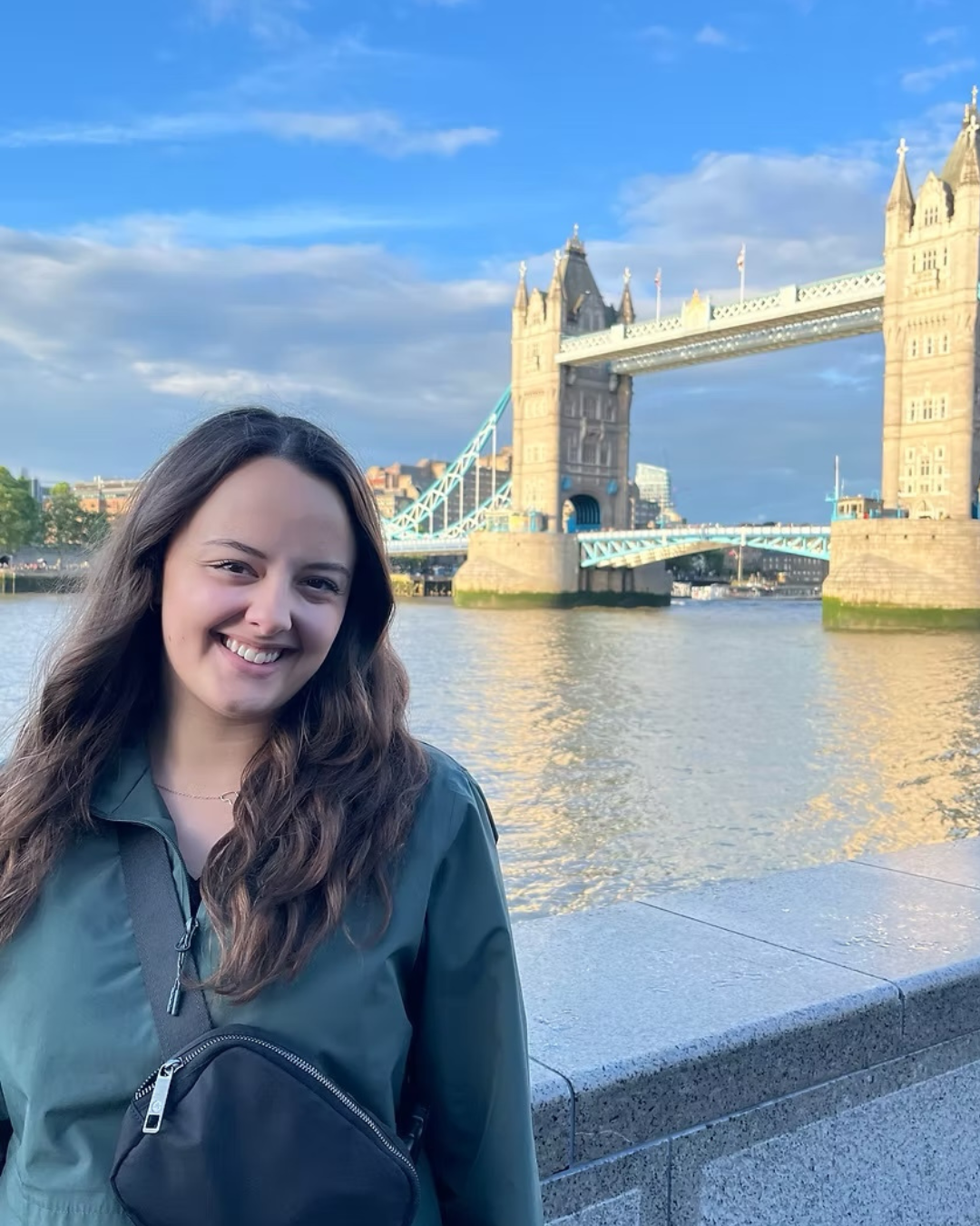 Smiling woman with long brown hair standing near the River Thames in London, with Tower Bridge in the background on a partly cloudy day.