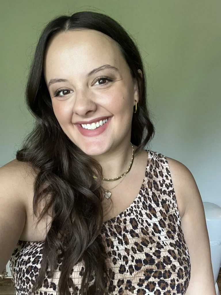 A woman with long, wavy dark brown hair smiling at the camera, wearing a leopard print sleeveless top, gold jewelry, and a green background.