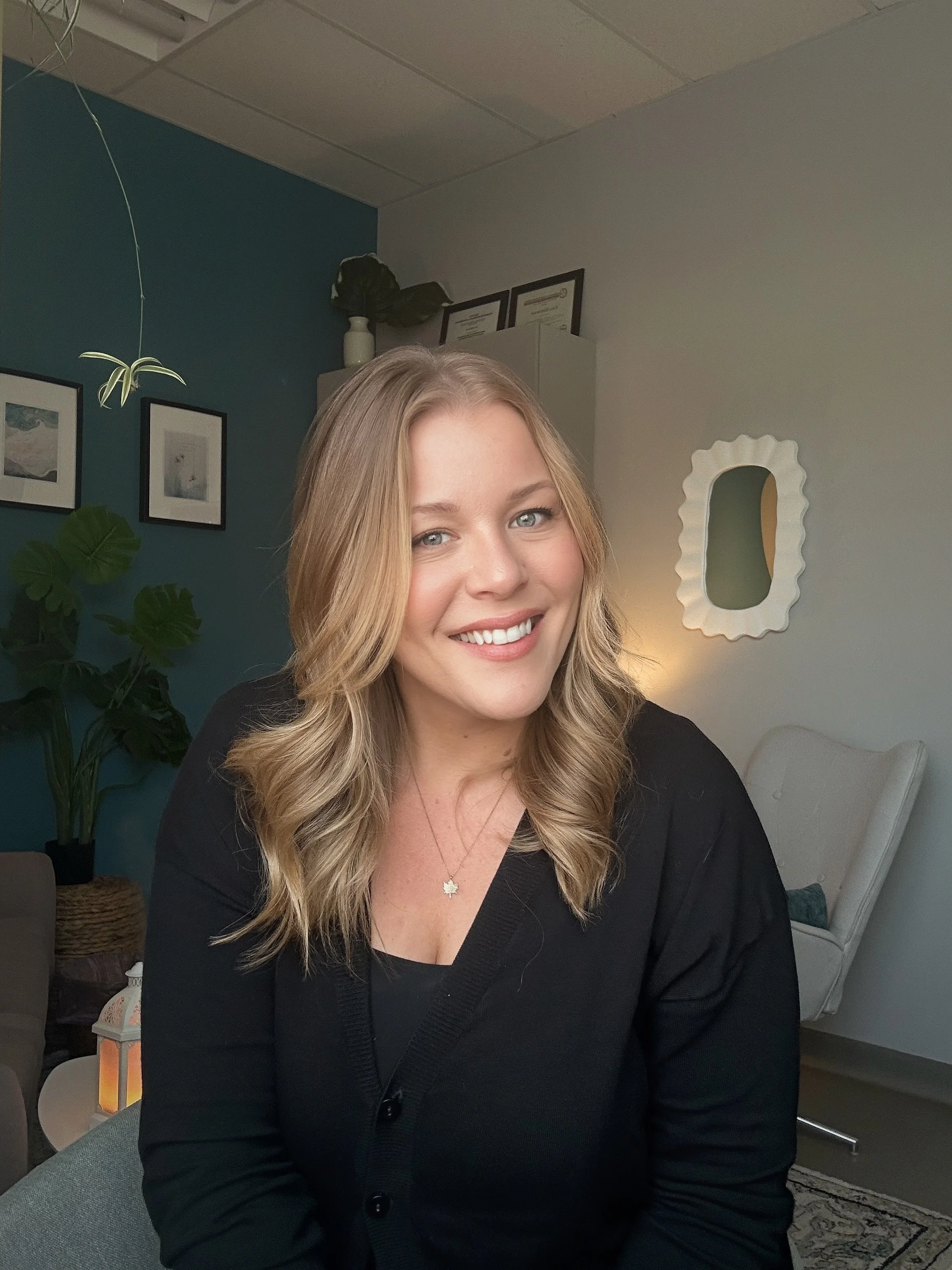 A woman with blonde, wavy hair smiles at the camera in a cozy room with framed pictures, plants, and a decorative mirror.