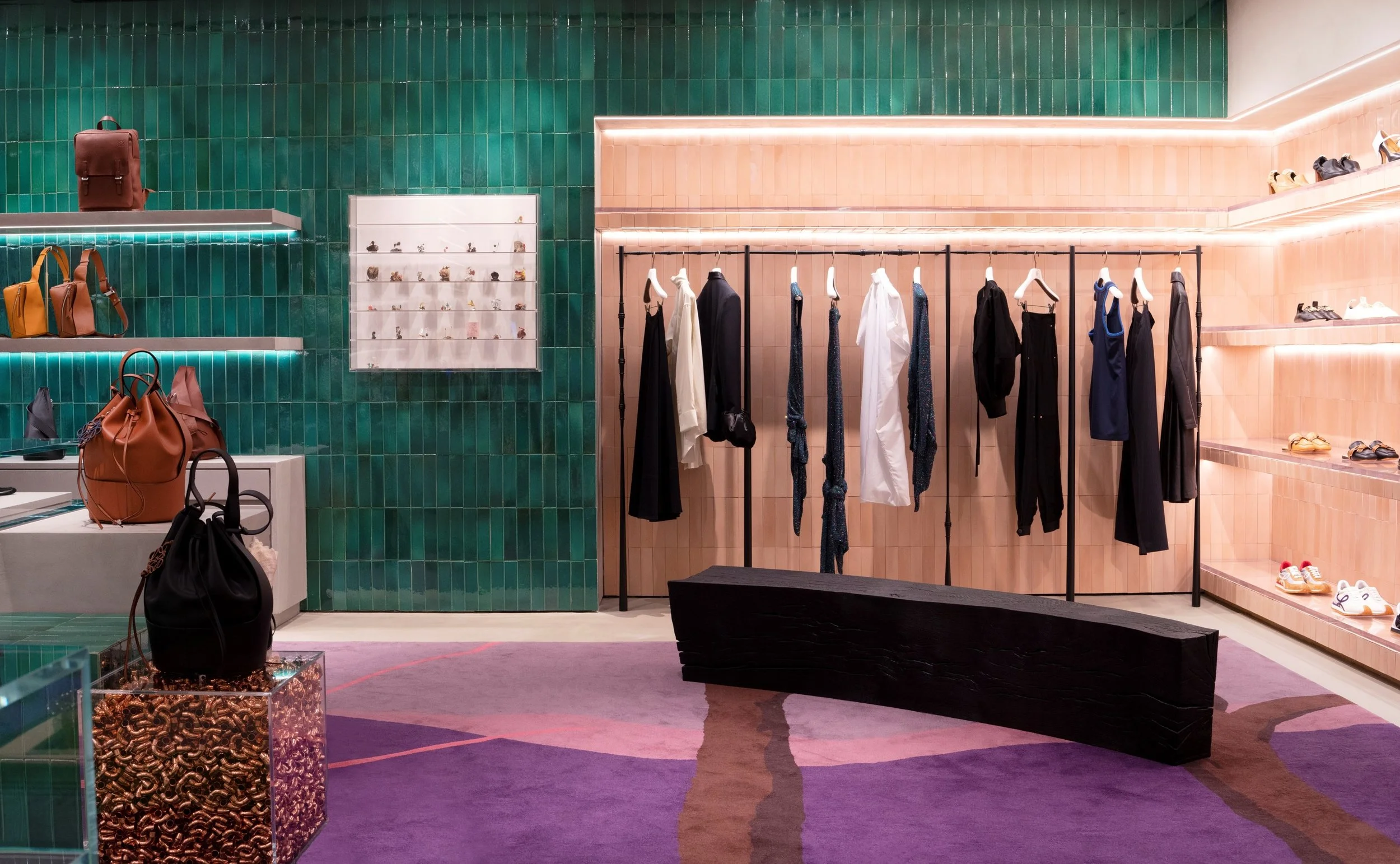 A retail clothing store displays dresses, shoes, and handbags against a pink and green tiled wall with shelving and clothing racks.