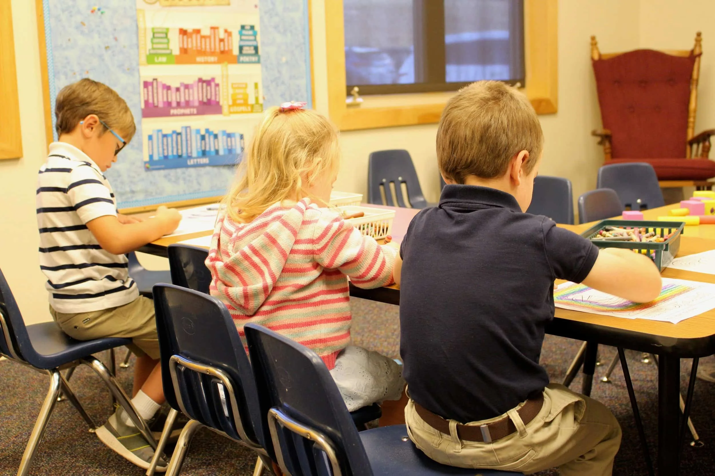 Three young children sit at desks in a classroom, coloring or writing with crayons and pencils.