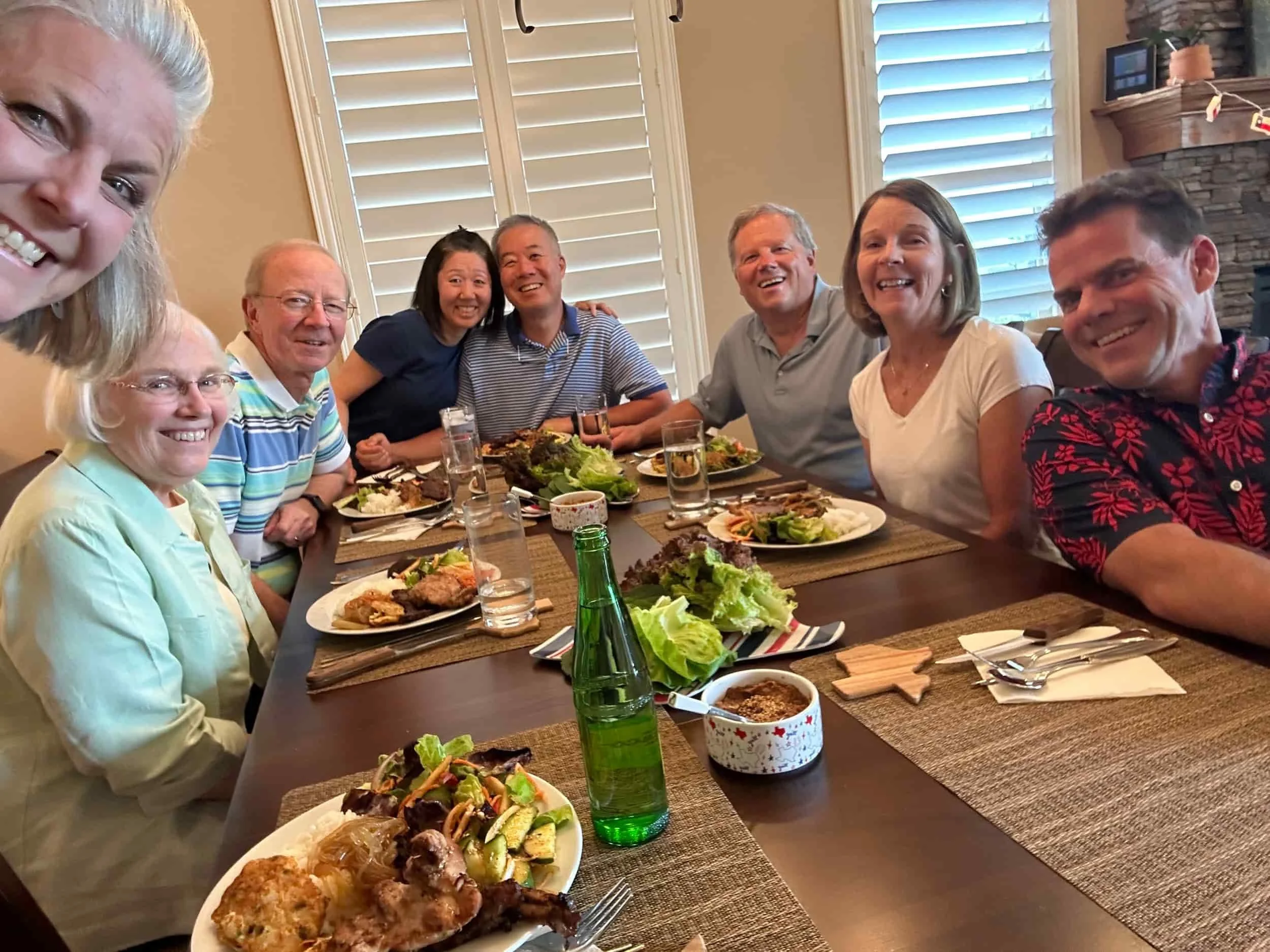 Eight smiling adults sit around a dining table enjoying a meal together, with plates of food and drinks in front of them.