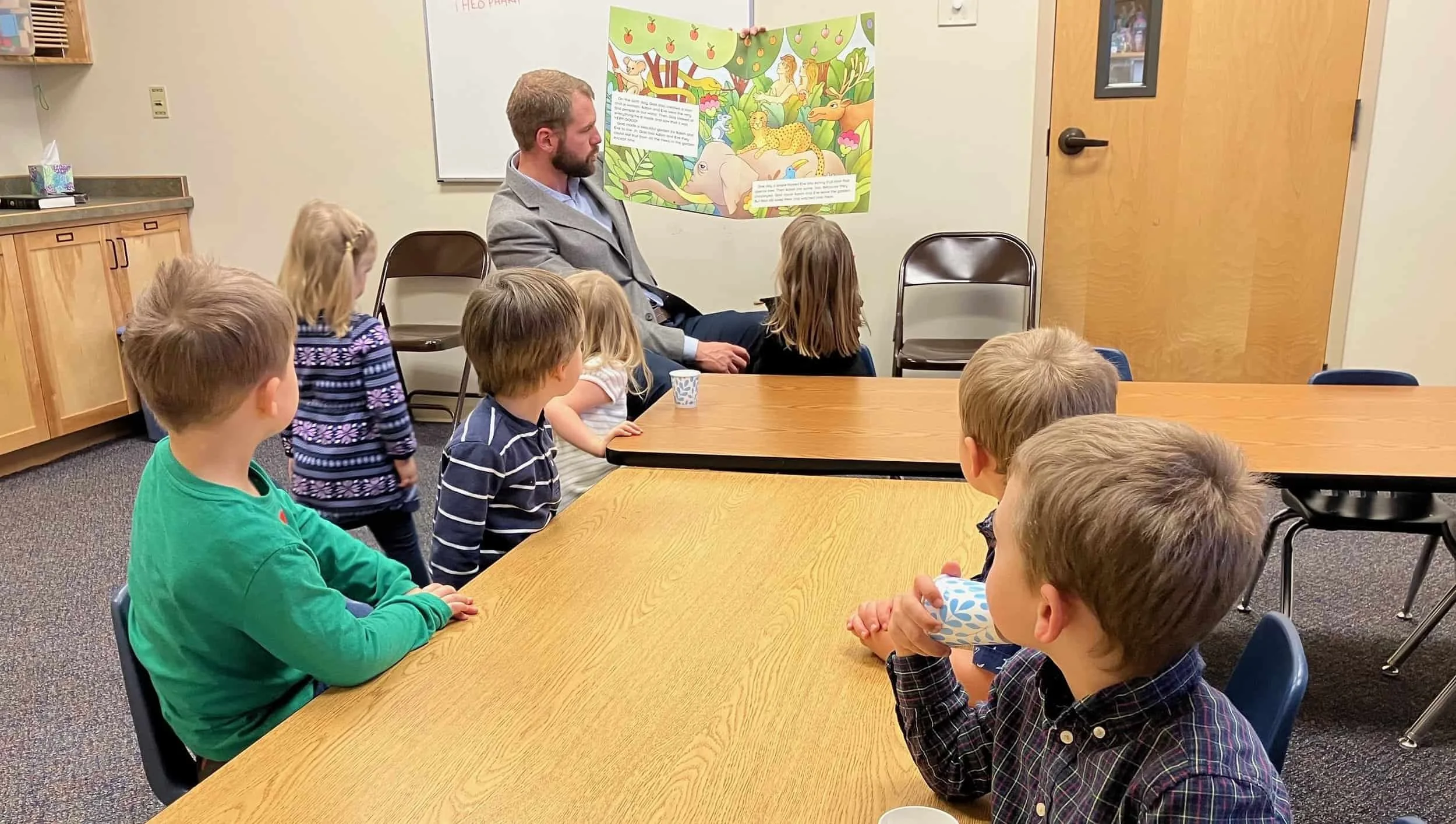 A man reads a picture book to a group of young children sitting around a table in a classroom.