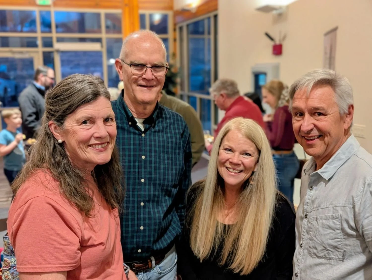 Four smiling adults pose together indoors at a social gathering, with other people blurred in the background.