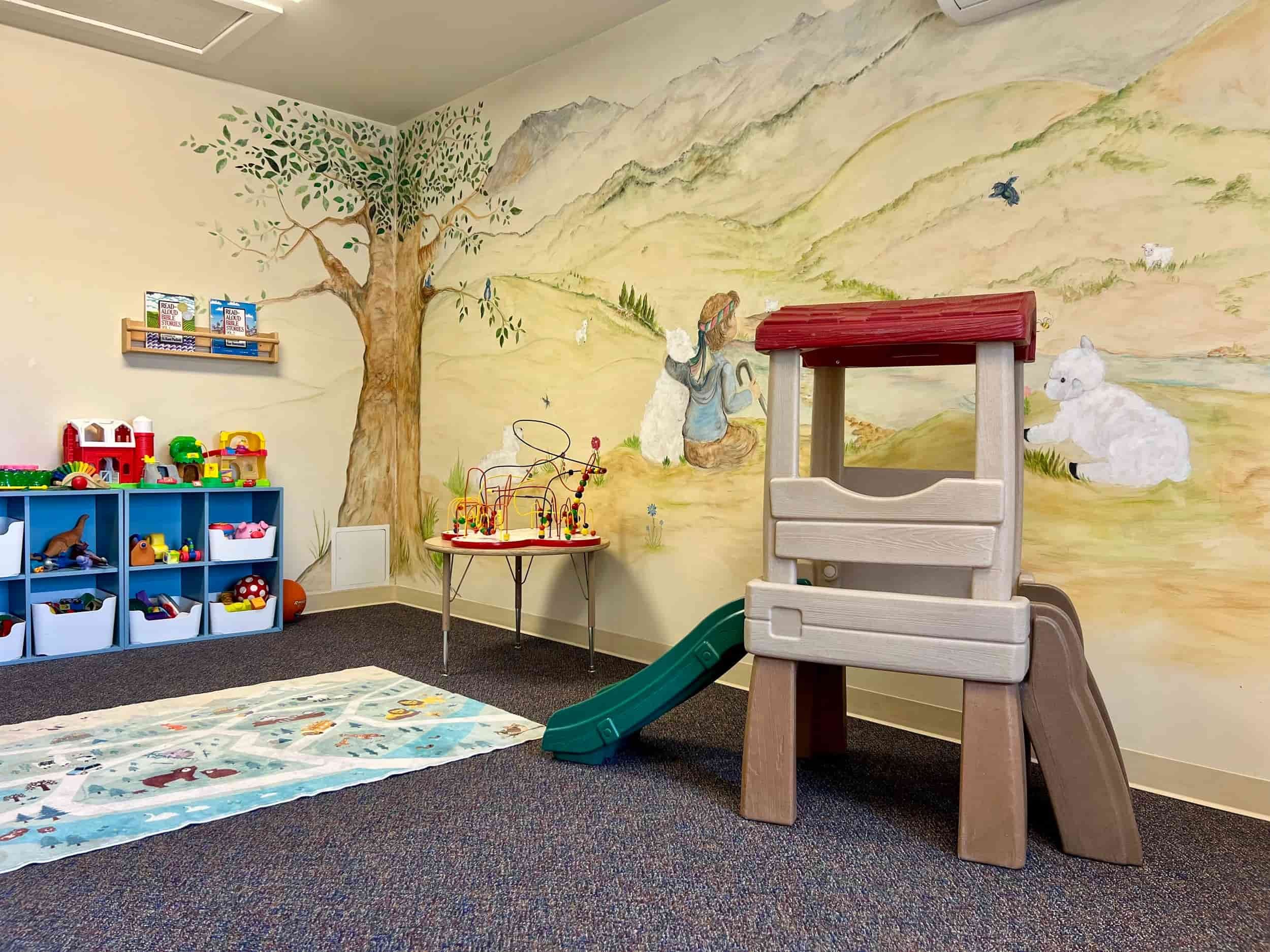 A colorful nursery playroom with toys, a play structure with a slide, shelves, and a mural of animals and a tree on the wall.