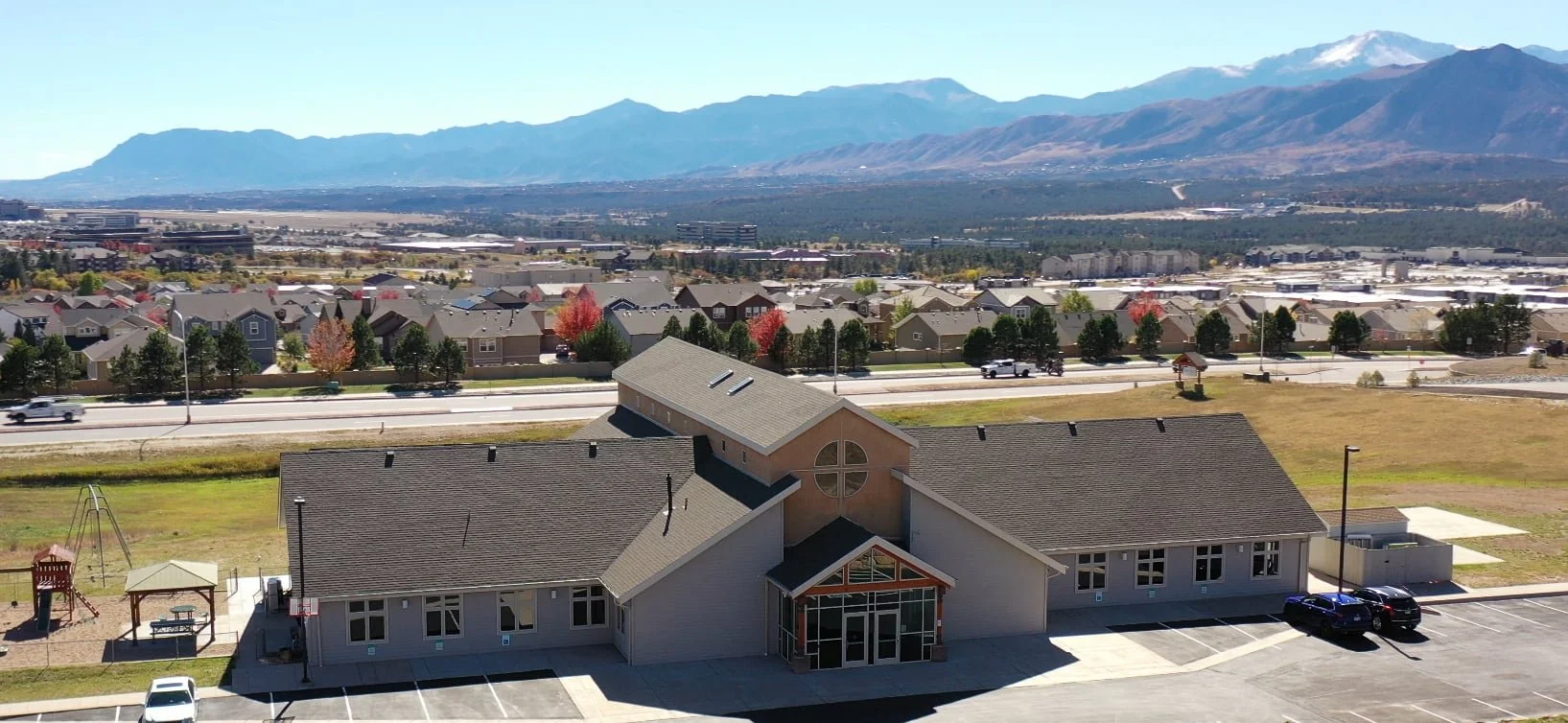 Forestgate Presbyterian Church, a large building with a peaked roof, stands before houses and a highway, mountains in the background.