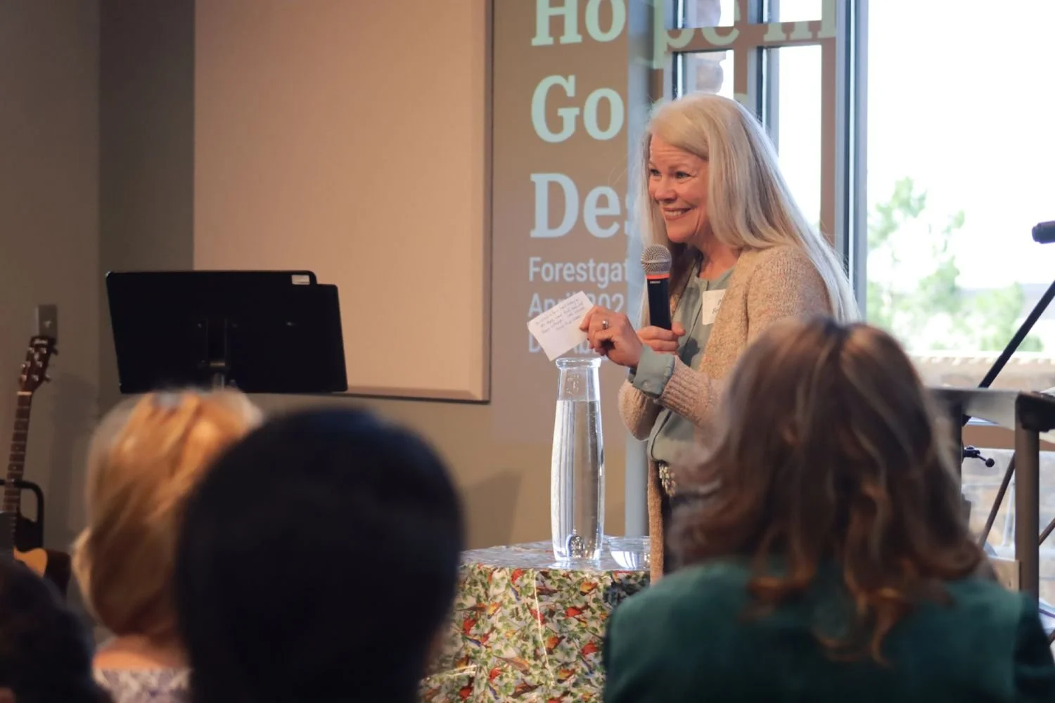 A woman speaks for Forestgate Women's Ministry, addressing an audience beside a table with a pitcher and papers.