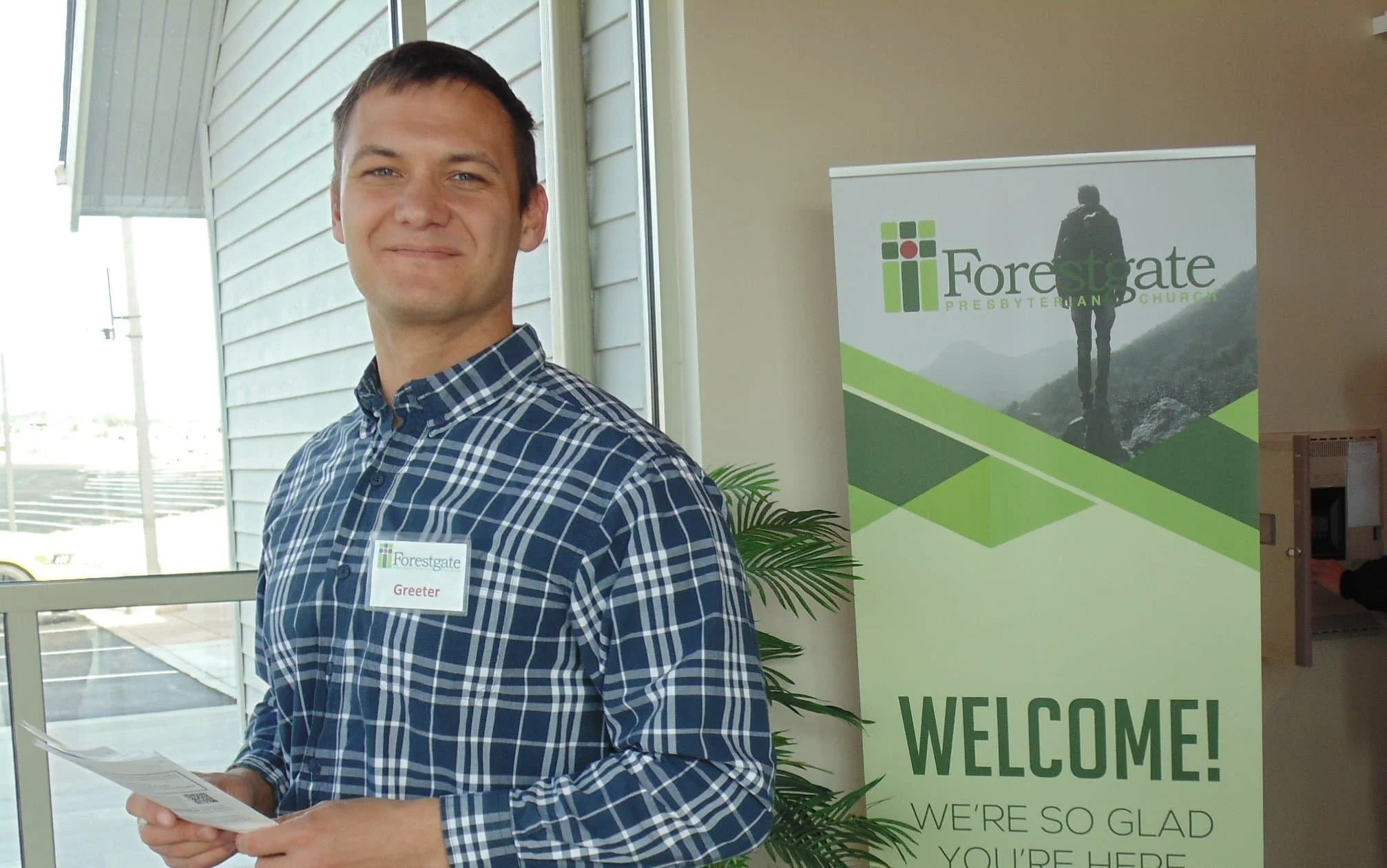 A man wearing a Greeter name tag smiles beside a Welcome! sign at Forestgate Presbyterian Church.