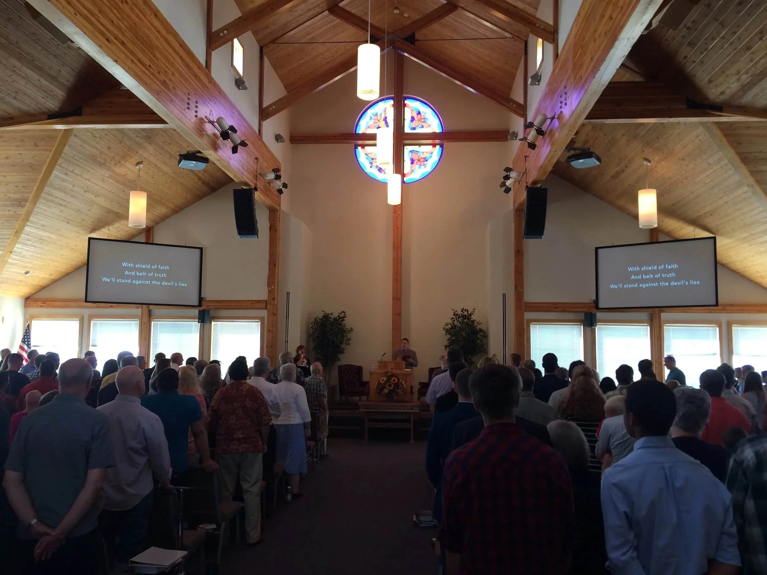 A congregation stands and faces forward in the Forestgate Presbyterian Church sanctuary, with wooden beams and a stained glass window above the altar area. Song lyrics are displayed on two screens.