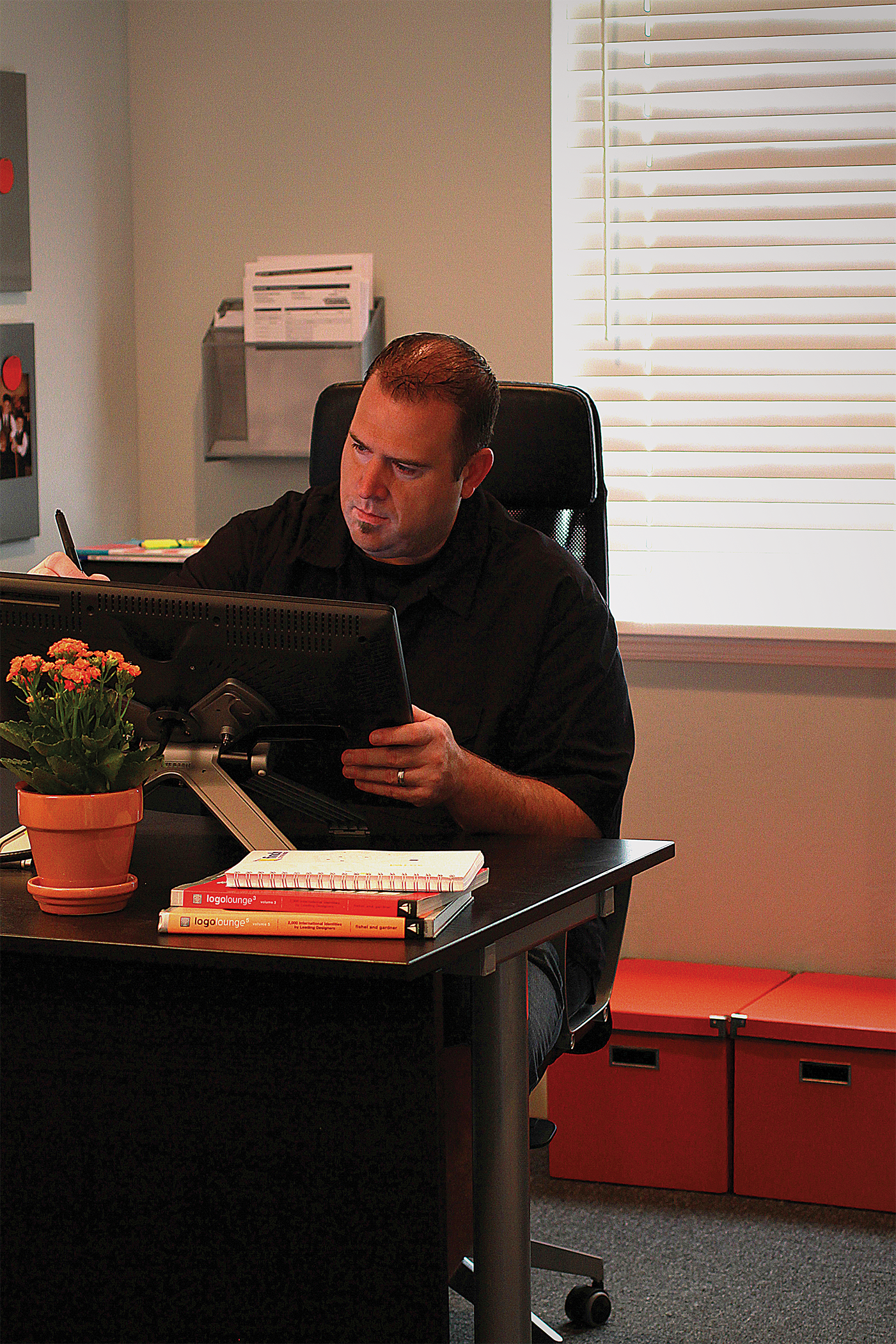 A man with short dark hair working at a black desk in an office, surrounded by books, a potted plant, a notepad, and a computer monitor, with blinds covering a window in the background.