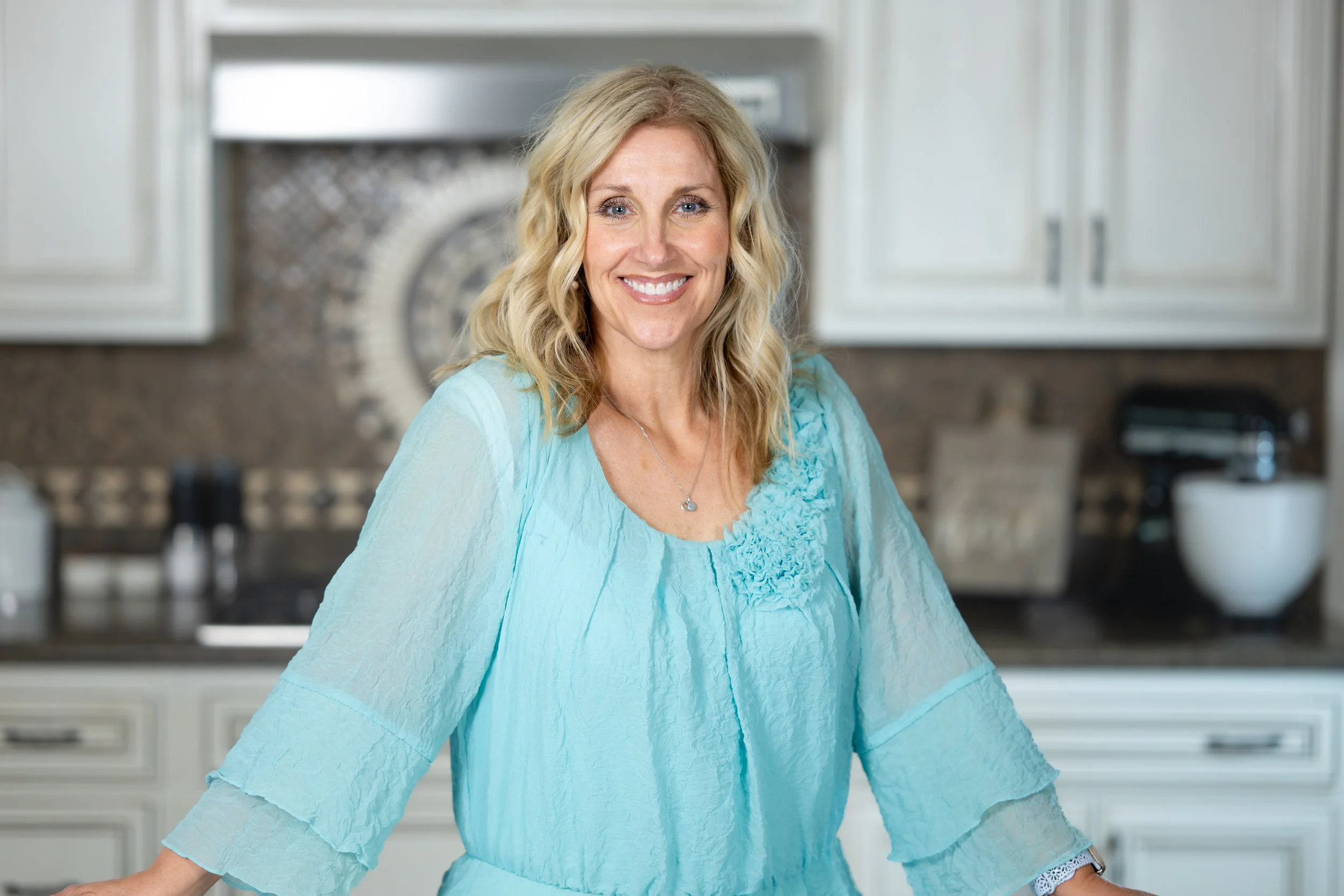 A woman with blonde hair is smiling and wearing a light blue blouse with ruffled details on the front, standing in a modern kitchen with white cabinets and a gray backsplash.