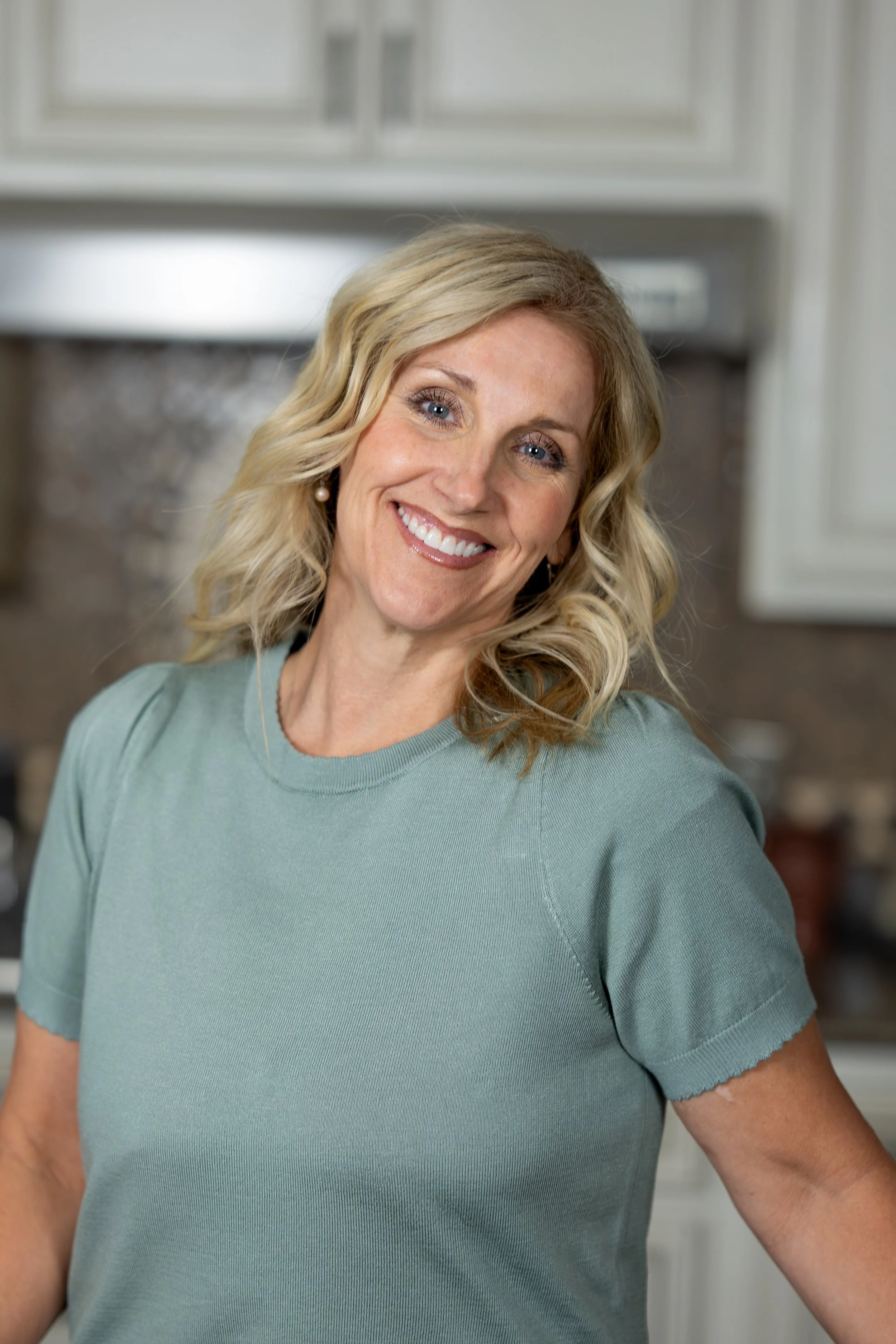 A woman with blonde wavy hair smiling and looking at the camera in a kitchen.