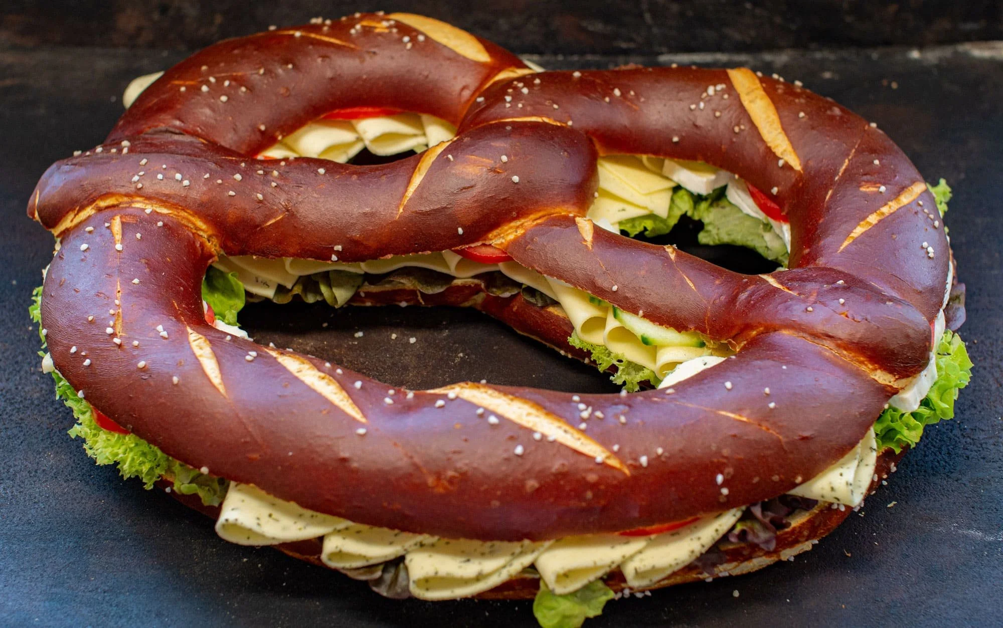 Ein große Laugenbrezel mit Sesam, belegt mit verschiedenem Käse, Wurst, Lachs, Salat, Tomaten und Gurken, auf einem schwarzen Untergrund.