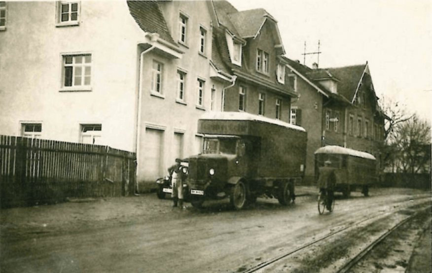 Schwarz-weiß-Foto zeigt eine Straße mit alten Lastwagen, einer Person mit Fahrrad und einem weiteren Mann, umgeben von mehrstöckigen alten Gebäuden.