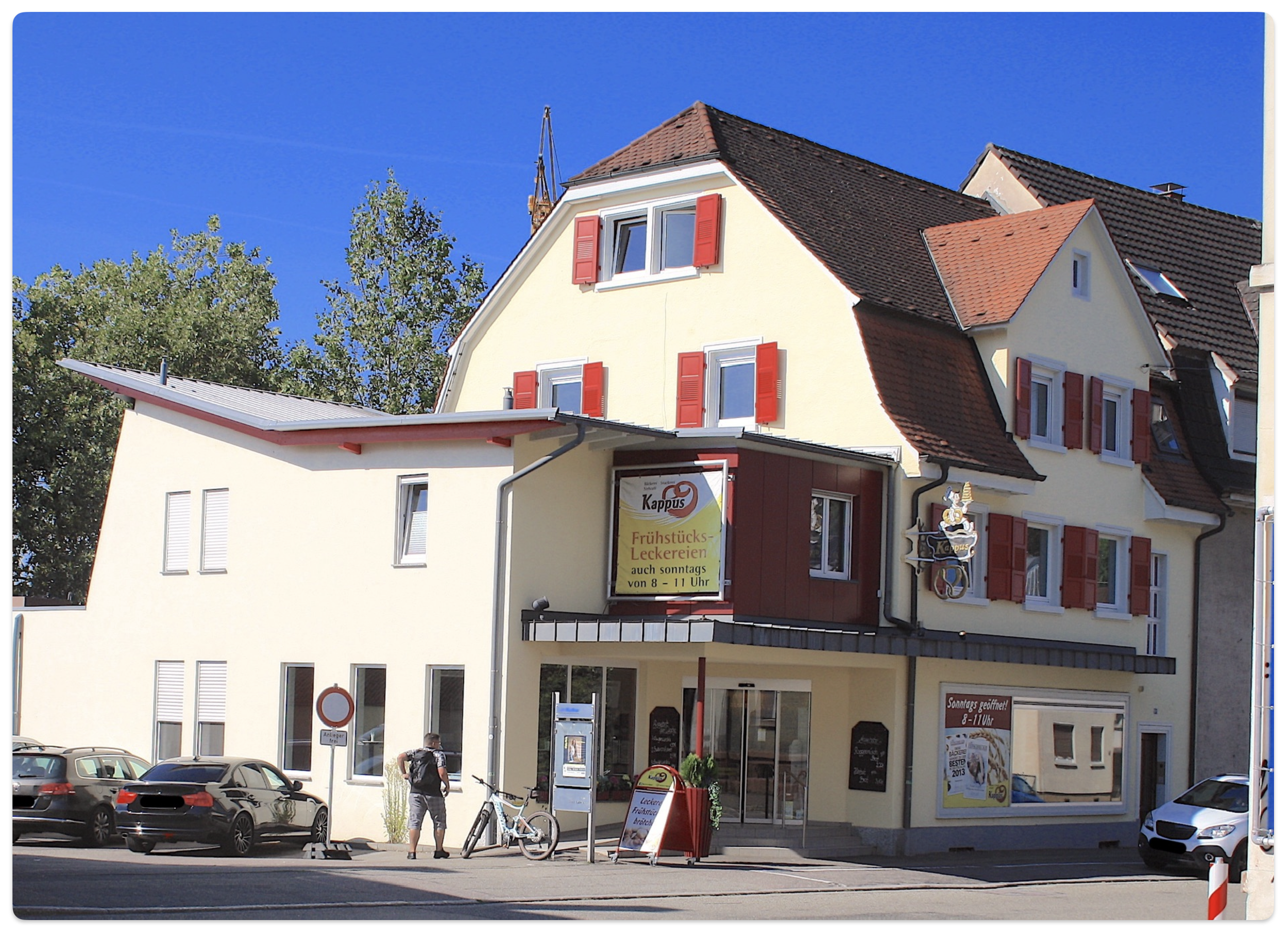 Ein mehrstöckiges Gebäude mit weißen Wänden und roten Fensterläden, Geschäfte im Erdgeschoss, Autos parken davor, eine Person steht mit einem Fahrrad auf dem Gehweg, blauer Himmel im Hintergrund.