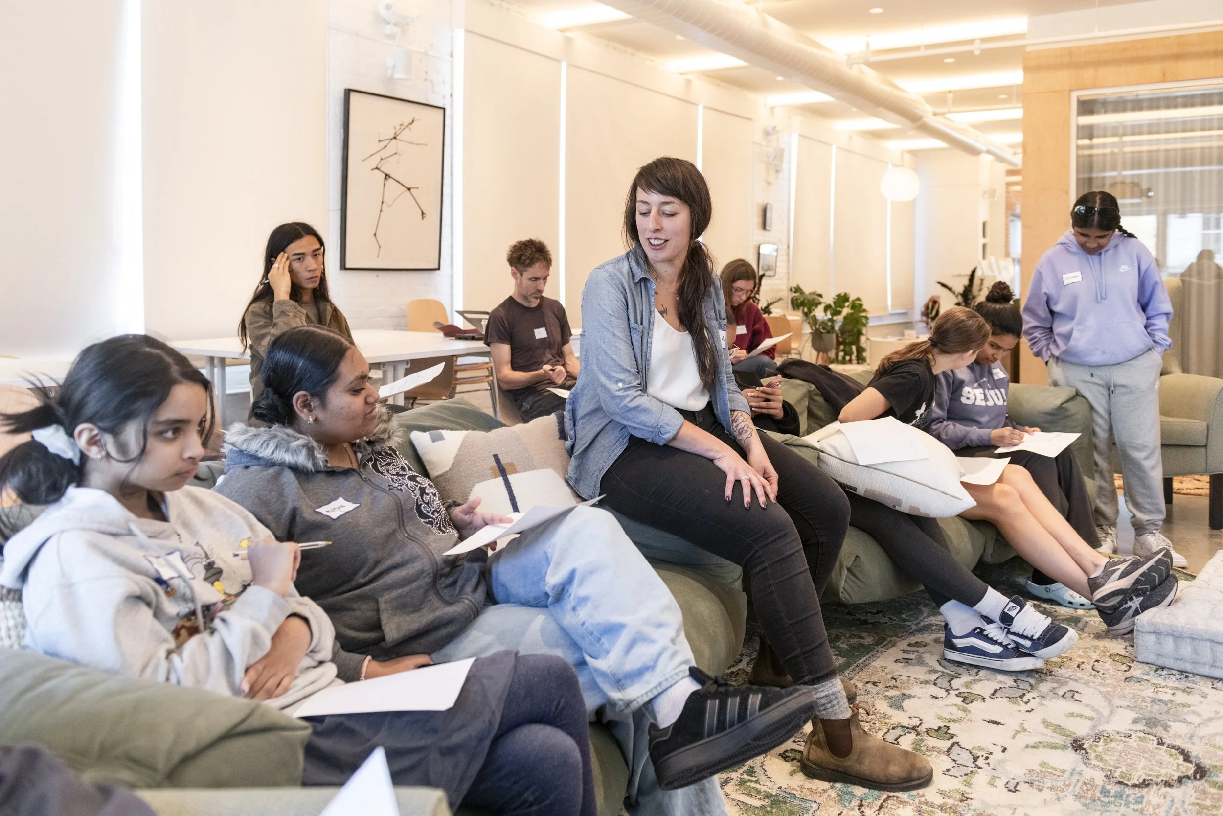 A group of young women and a woman sitting on a couch in a well-lit modern room, some taking notes, engaged in conversation or listening attentively.