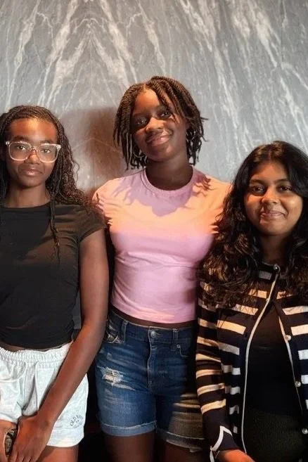 Three young girls posing together against a marbled wall. The girl on the left wears glasses, a black T-shirt, and white shorts. The girl in the middle wears a pink T-shirt and denim shorts. The girl on the right has curly hair, a striped jacket, and a dark top.