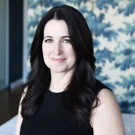 A woman with long black hair smiling in front of a window with a view of trees.