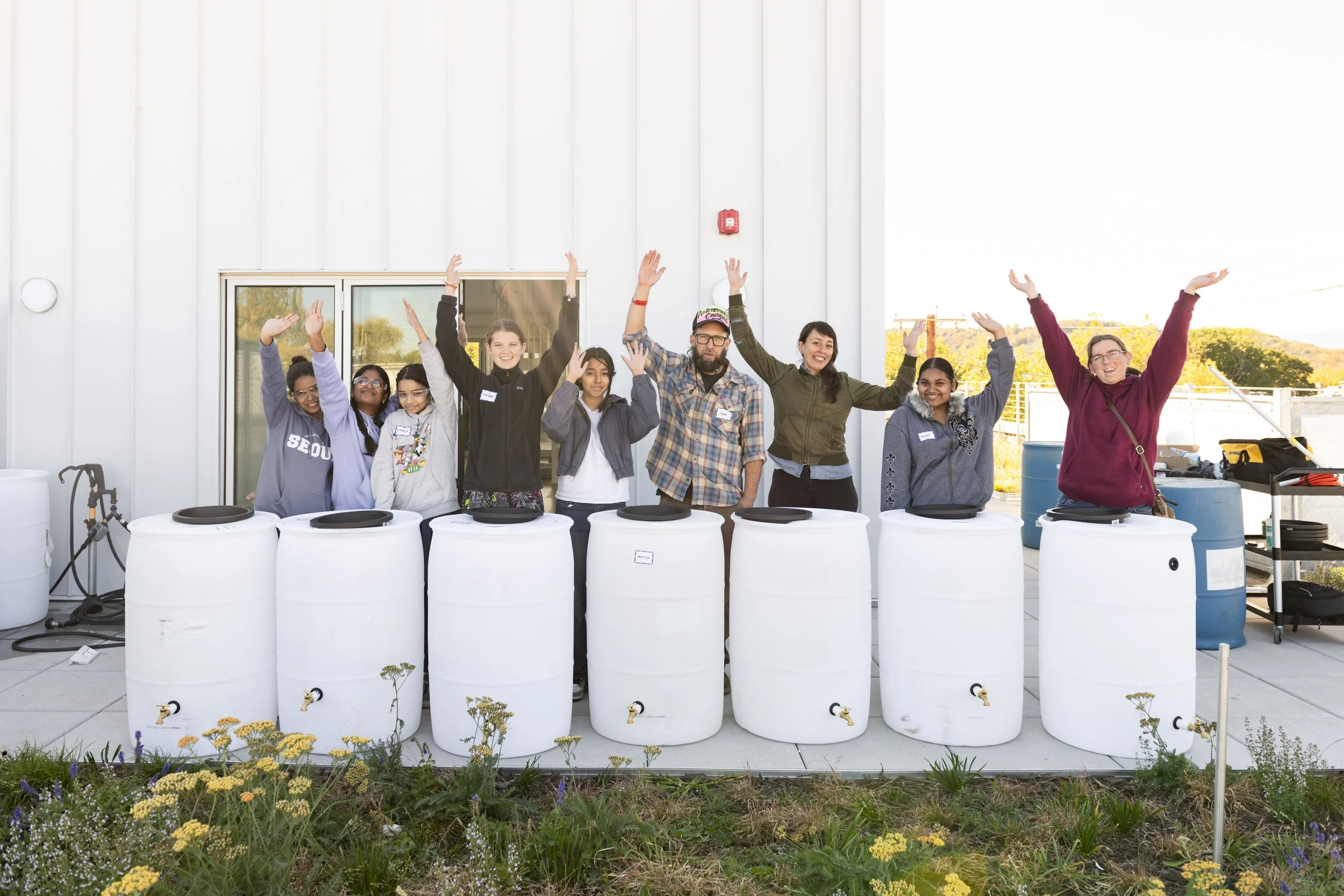 Group of eight people standing behind white rain barrels with spigots, smiling and raising their arms in celebration outside a building.