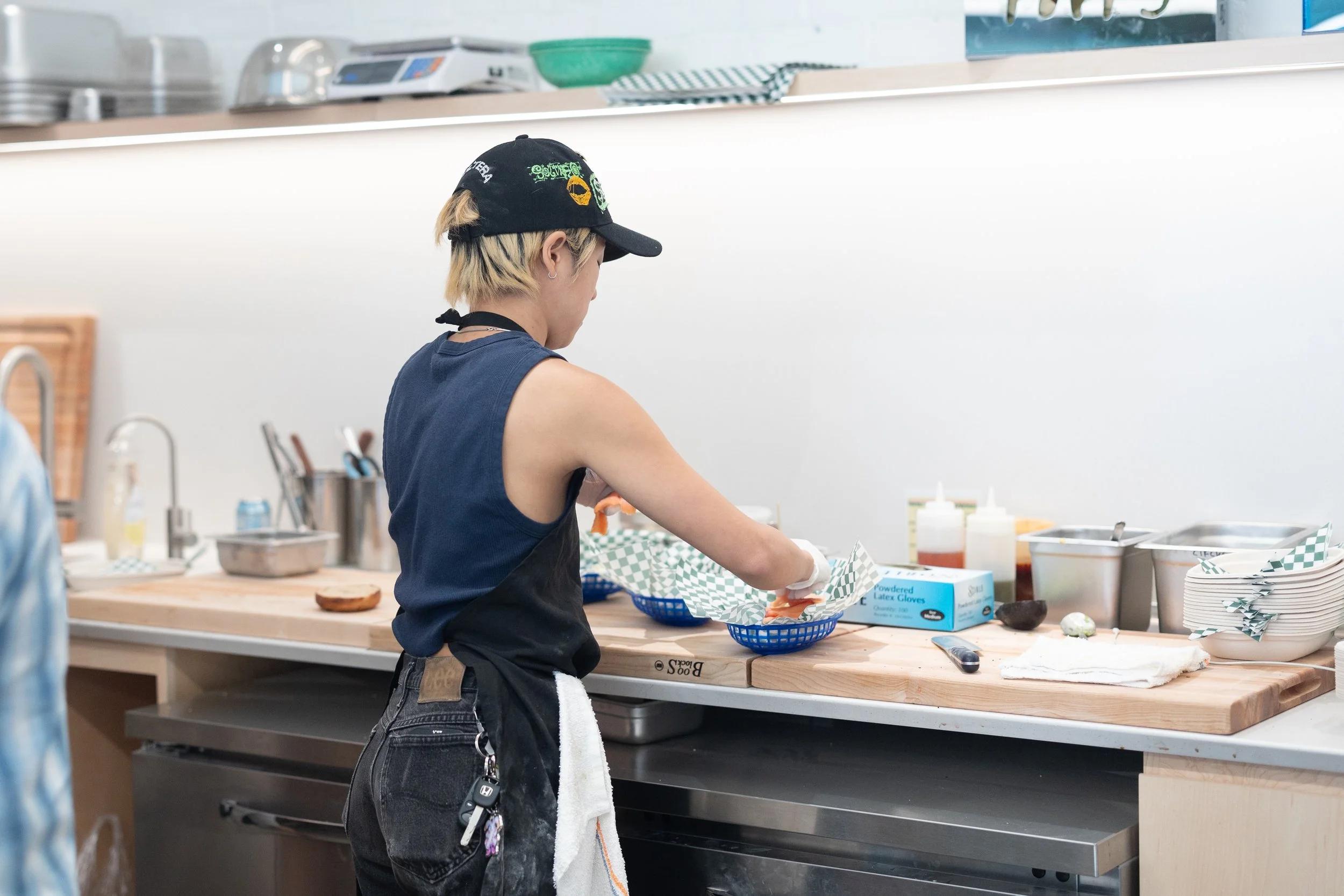 A young woman with short blonde hair wearing a black baseball cap, a sleeveless navy shirt, and black pants prepares food wearing gloves in a restaurant kitchen.