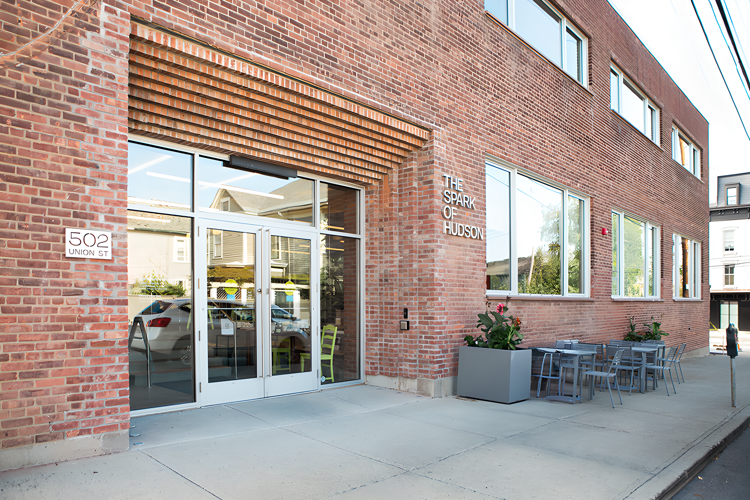 Exterior view of a red brick building with large windows and a sign that reads 'THE SPARK OF HUDSON.' There is an entrance with glass double doors, a sidewalk with outdoor seating, and potted plants with flowers.