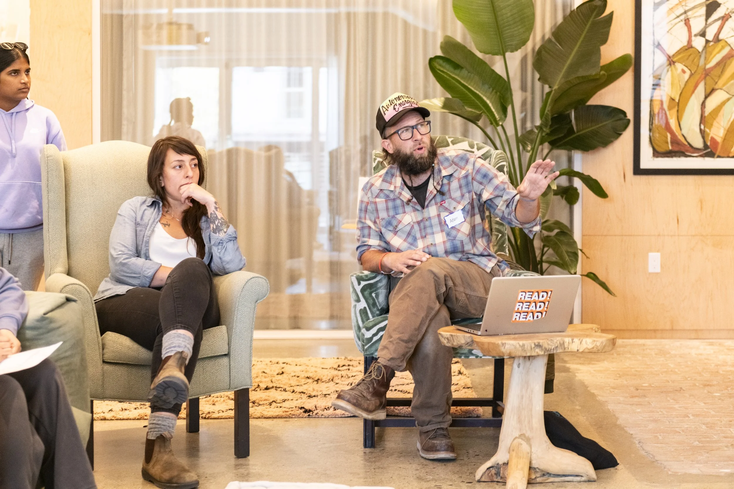 A group of people in a living room setting, a man with glasses, a beard, and a plaid shirt is speaking and gesturing with his hand, sitting in a patterned chair. Others are listening, including a woman seated on a green armchair and a person standing nearby. A laptop on a wooden table with a 'READ! READ! READ!' sticker, a large leafy plant, and colorful artwork on the wall are also visible.