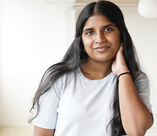 A young woman with long black hair and brown skin, wearing a light gray t-shirt, looks at the camera with a slight smile, holding her neck gently with her right hand.