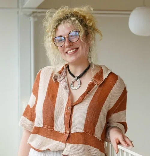 A woman with curly blonde hair and glasses smiling while leaning on a white railing indoors, wearing a striped brown and beige shirt and a black necklace.