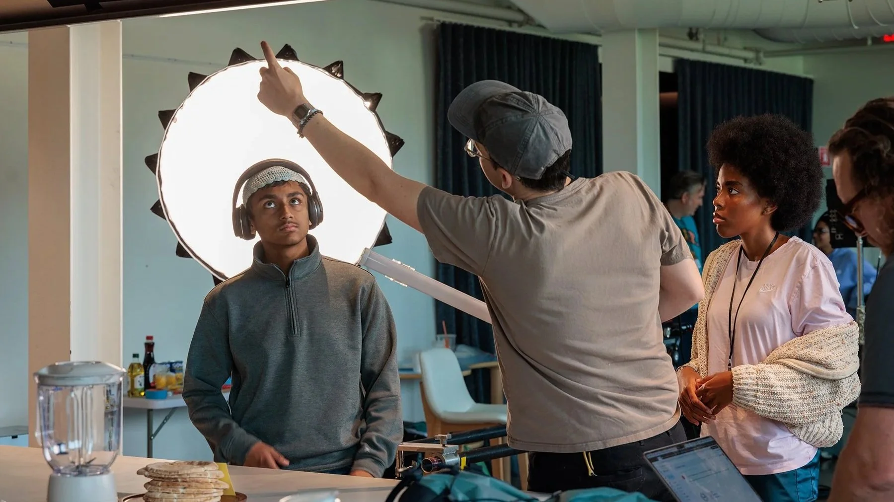 A young boy wearing headphones and a gray jacket is being filmed or photographed, with a bright reflector and a person adjusting it. Two women are watching, one with curly hair and a light pink shirt, and the other with glasses. A table with some food and a laptop is also visible.
