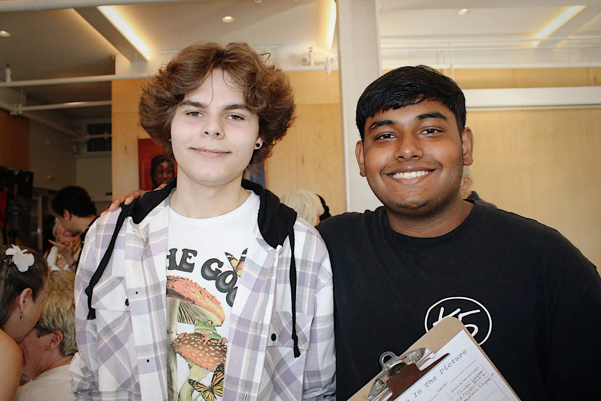Two smiling young men, one with light hair and one with dark hair, standing indoors with their arms around each other's shoulders. The young man on the right is holding a clipboard.